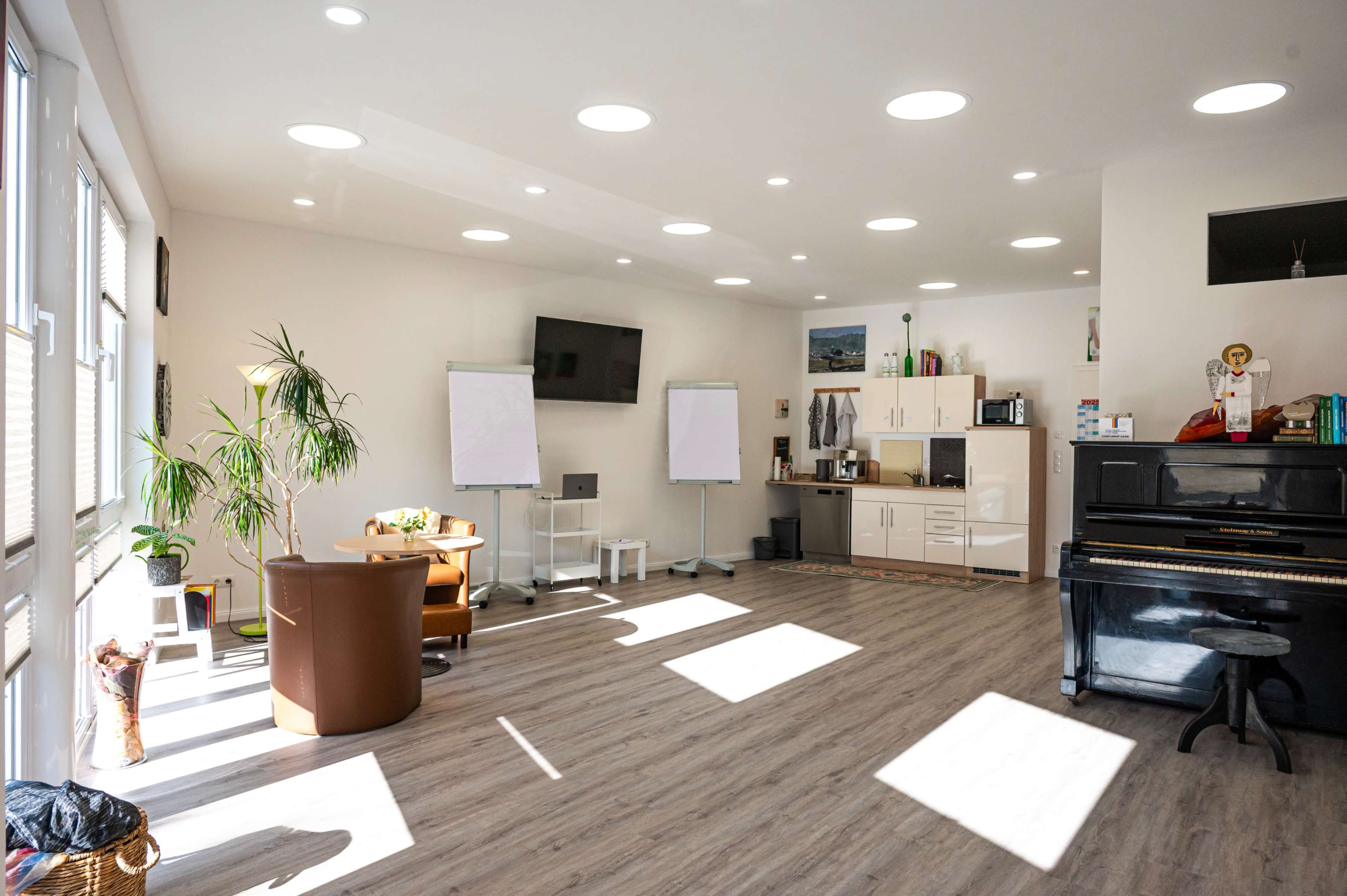 The image shows a bright, spacious room with wooden flooring, featuring a piano, a kitchenette, two whiteboards, and a seating area with a chair and a small table.