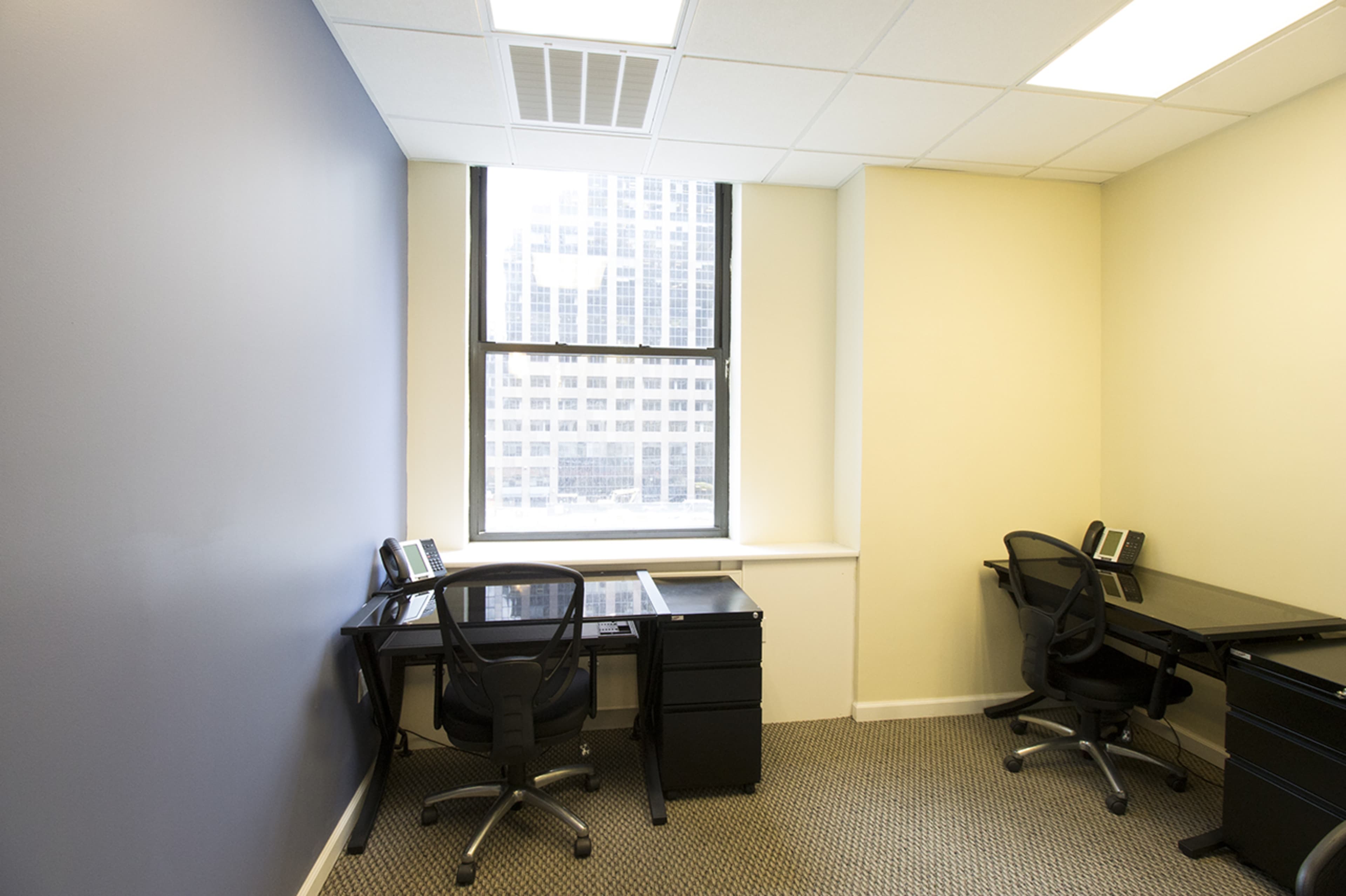 A small office space with two black desks and chairs, separated by a window overlooking a cityscape.