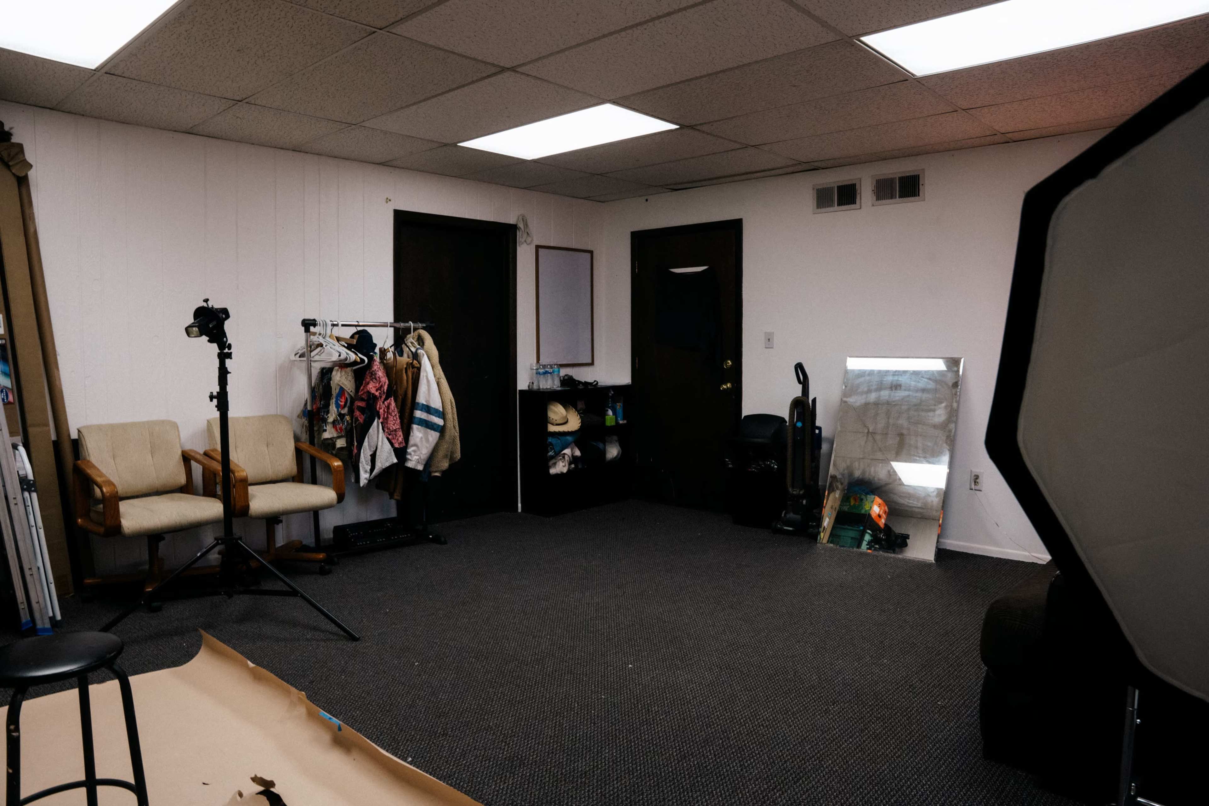 The image depicts a vacant room featuring a clothing rack, a camera stand, a large mirror, and two chairs, with minimal decorations on the wall.