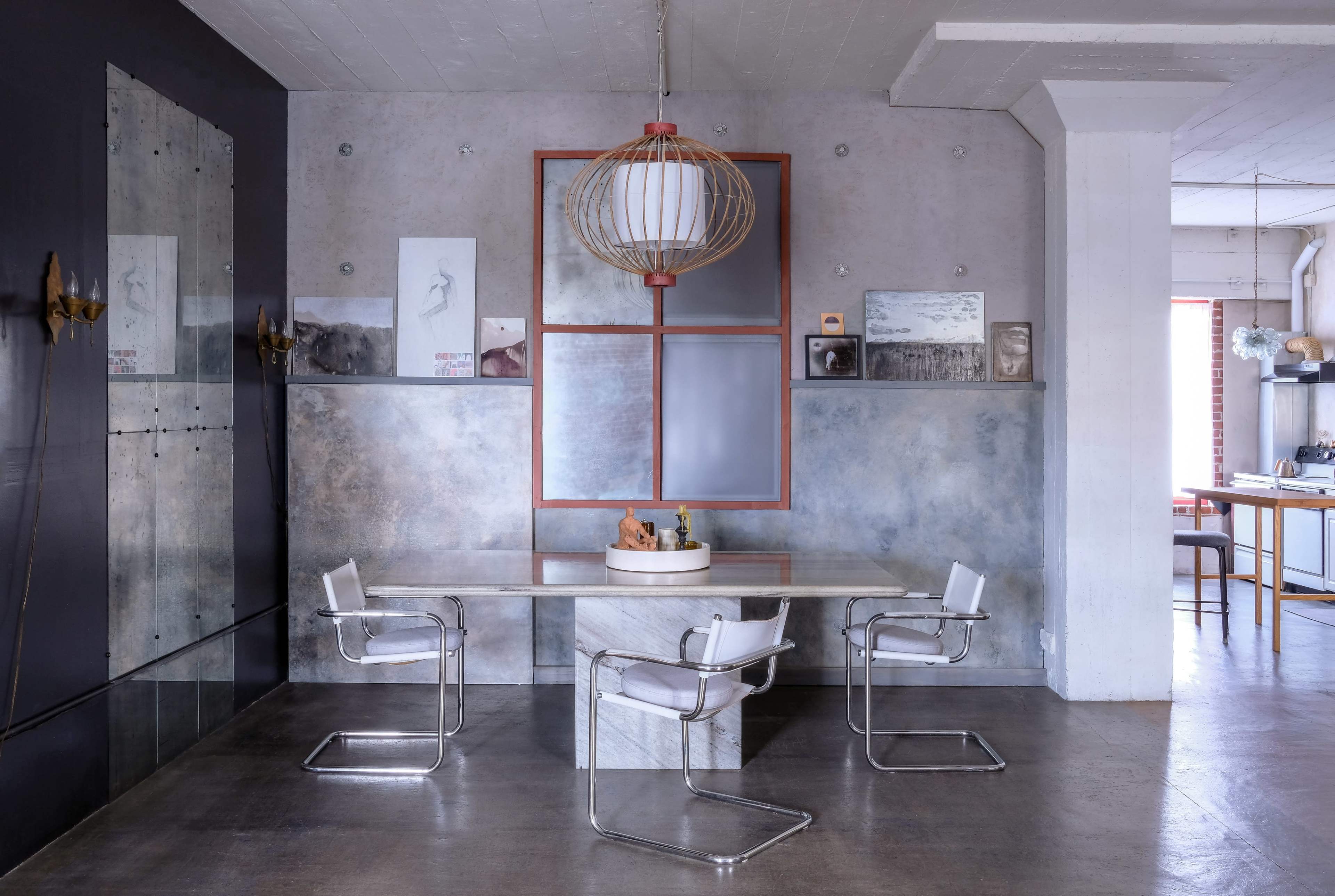 A modern dining area features a stone table surrounded by four metal-framed chairs, with a large pendant light hanging above and artwork displayed on the walls.