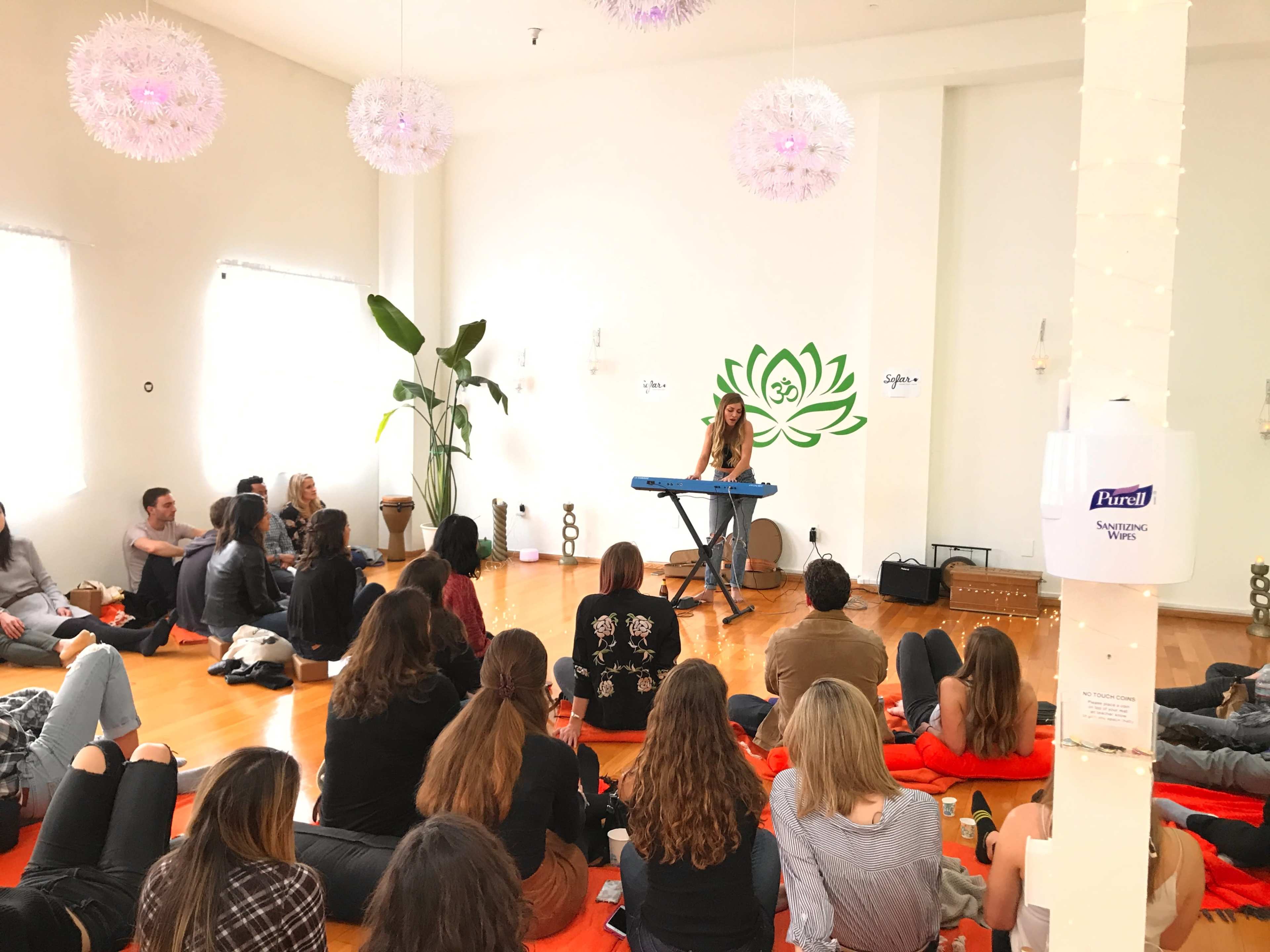 A woman performs at a keyboard for an audience seated on the floor in a warmly lit room decorated with plants and wall art.