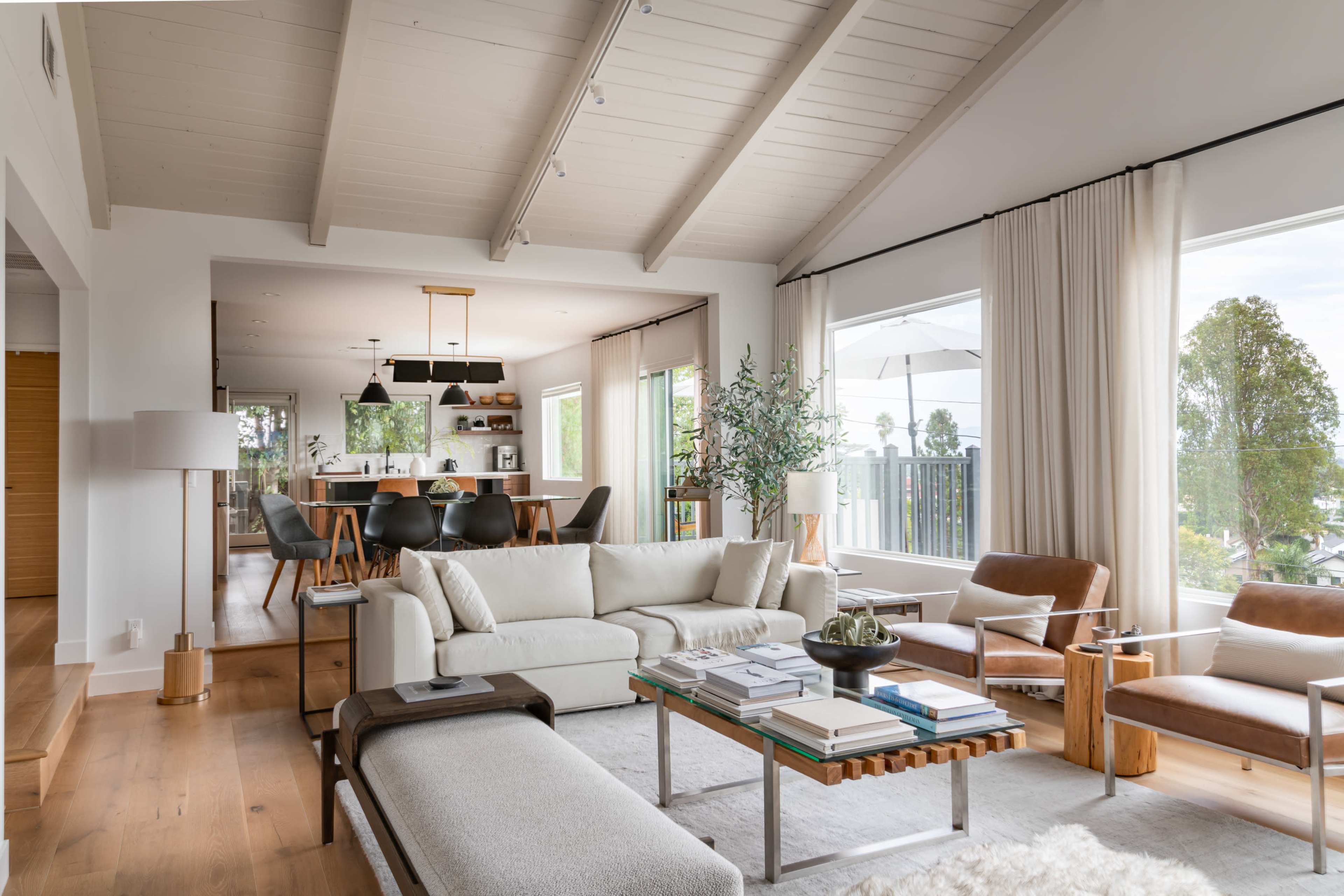 A bright living room features a large white sofa, two leather chairs, and a wooden coffee table, with a view of a kitchen and outdoor patio through large windows.