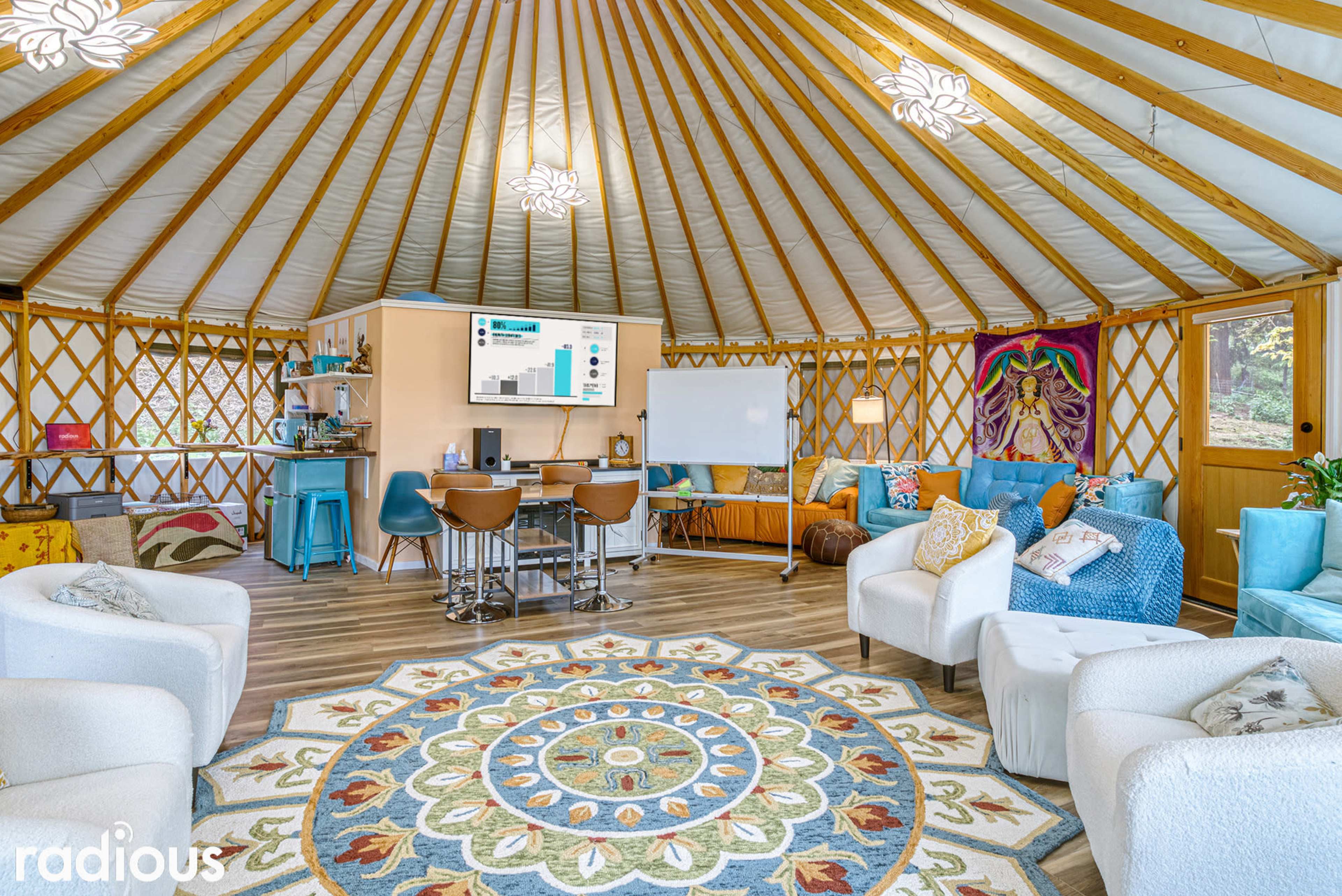 The interior of a yurt, featuring a circular seating area, a decorative rug, and a presentation screen against a backdrop of wooden lattice walls.