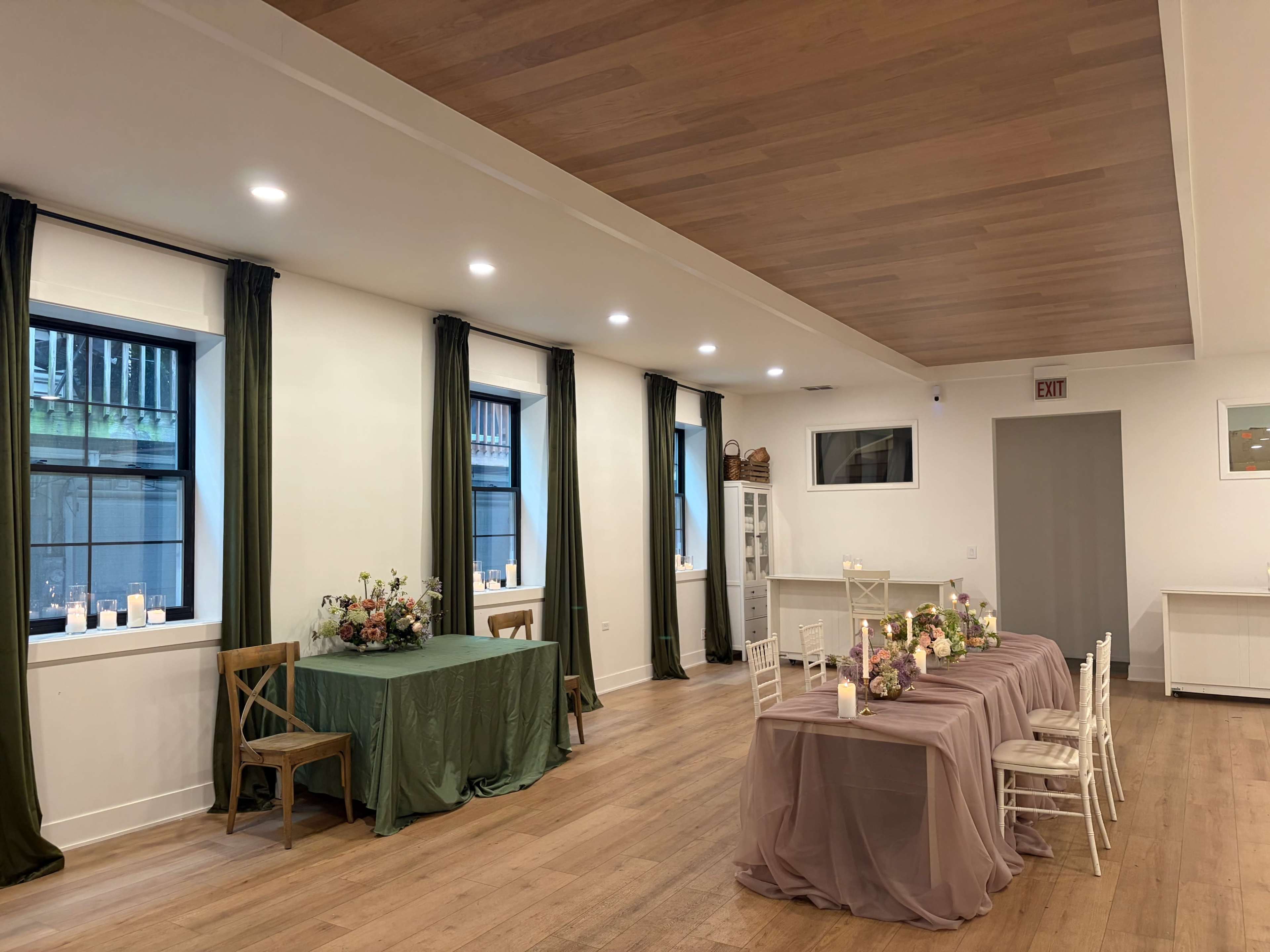 The image showcases a spacious indoor setting with two tables adorned with tablecloths, candles, and floral centerpieces, surrounded by white chairs and large windows.
