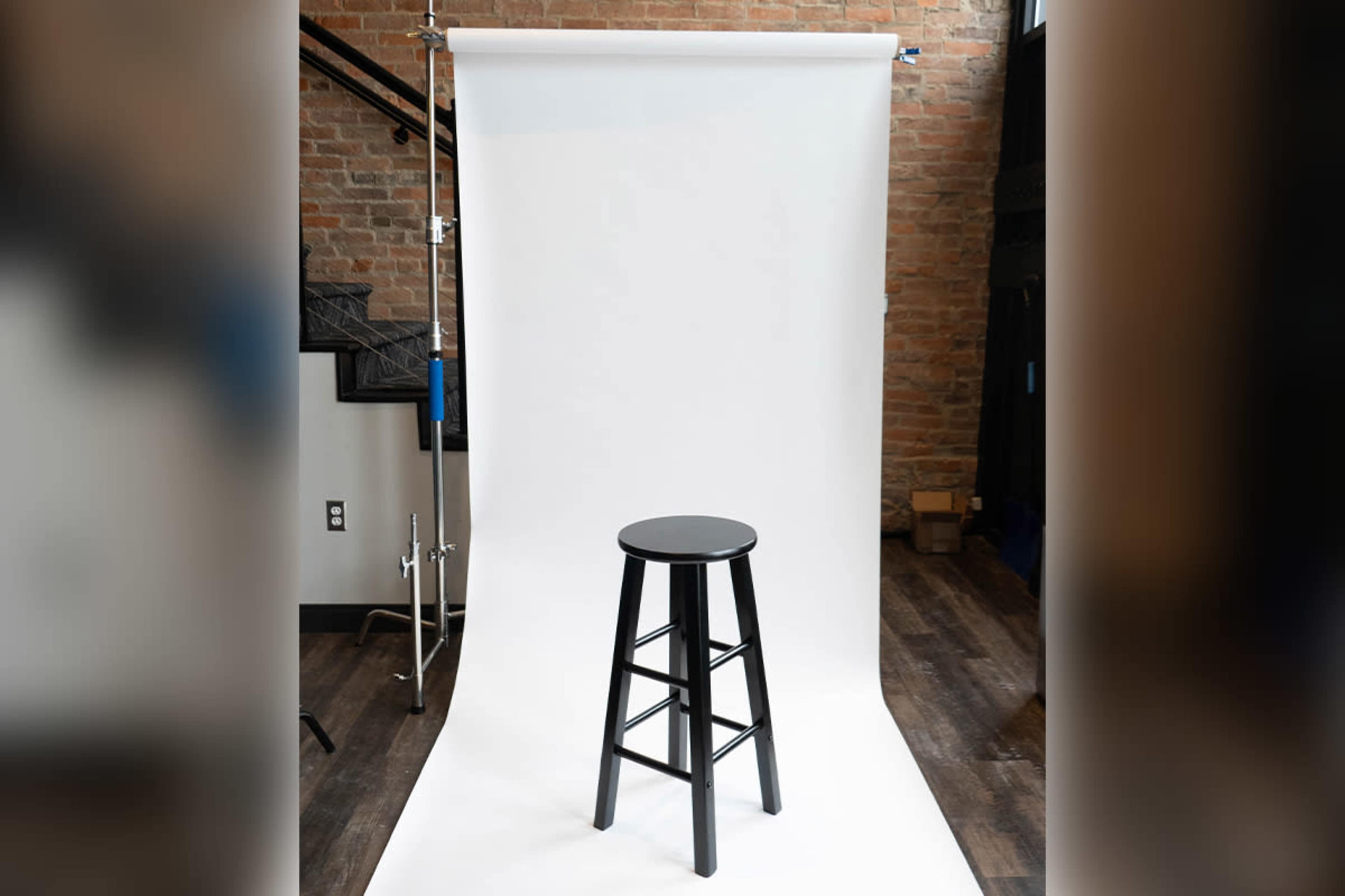 A black stool is positioned in front of a white backdrop in a spacious studio with exposed brick walls.