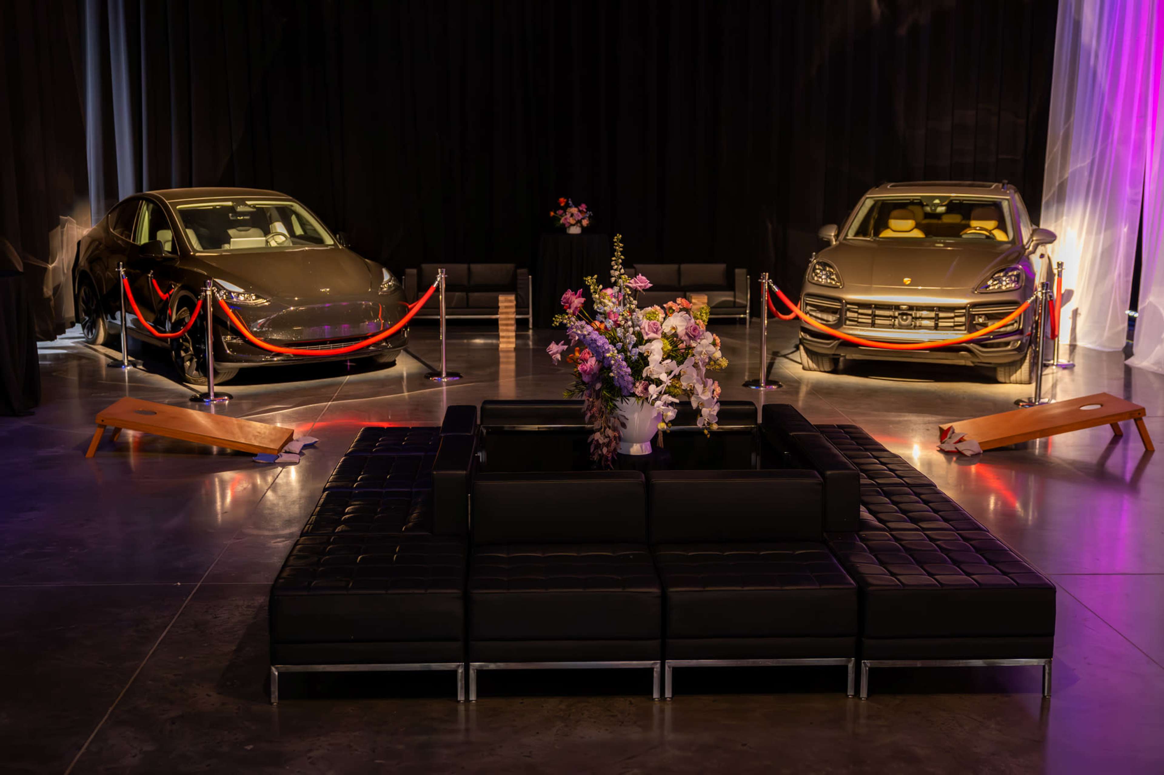 Two luxury cars are displayed on either side of a flower arrangement in a showcase area with black curtains and seating.