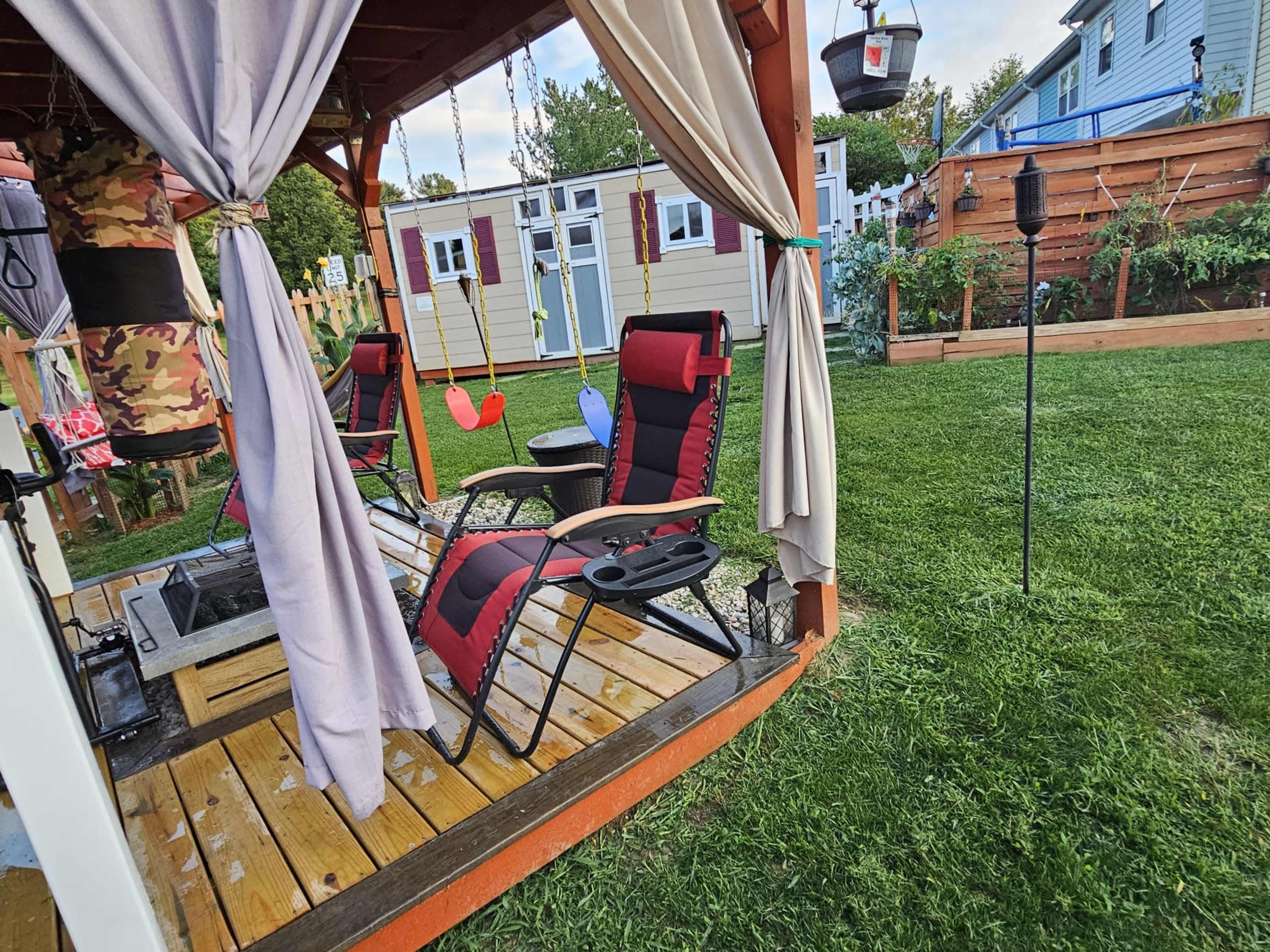 A gazebo with two reclining chairs and a small table is situated on a wooden deck in a backyard, surrounded by green grass and a vegetable garden.