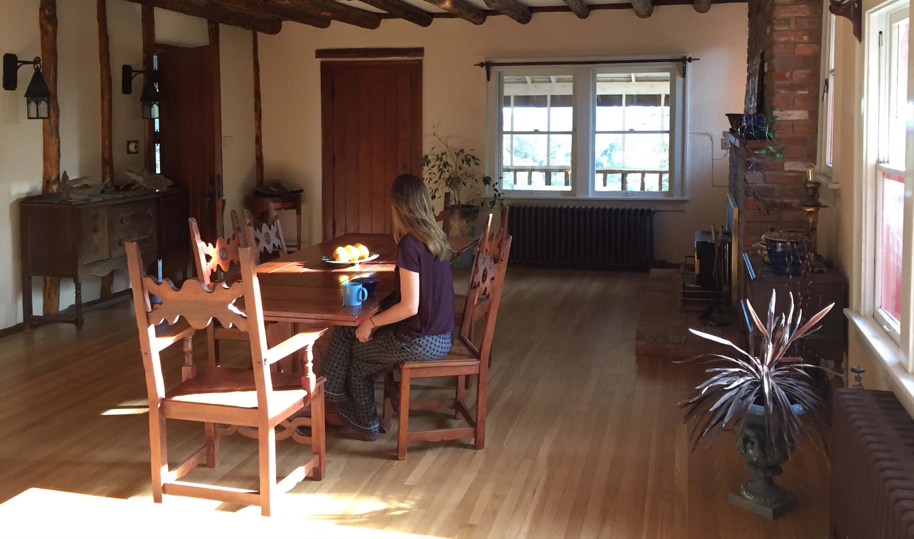 A woman sits at a wooden table in a spacious room with large windows and wooden beams on the ceiling.
