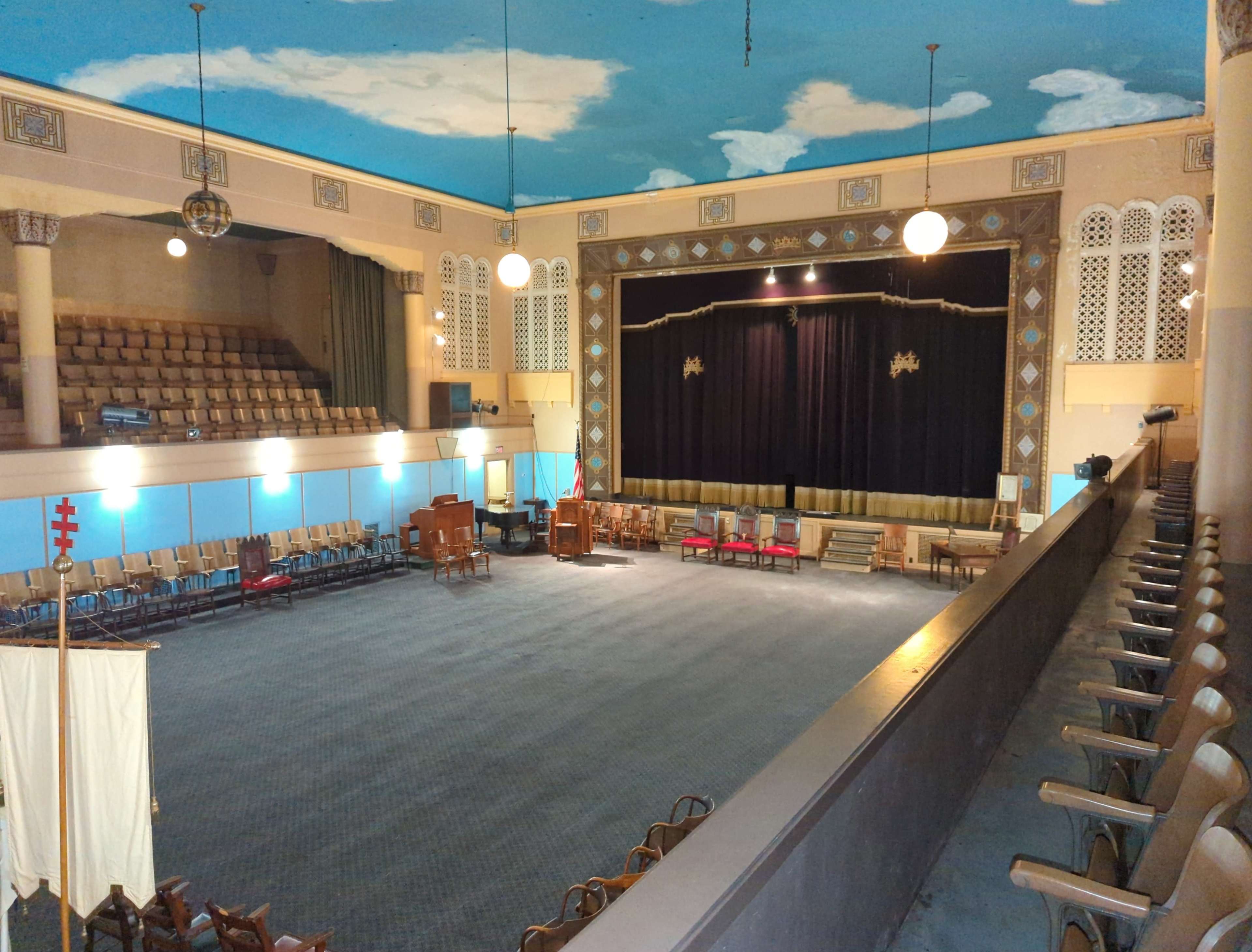 A spacious auditorium with a dark stage, featuring rows of empty chairs and a ceiling painted with clouds.