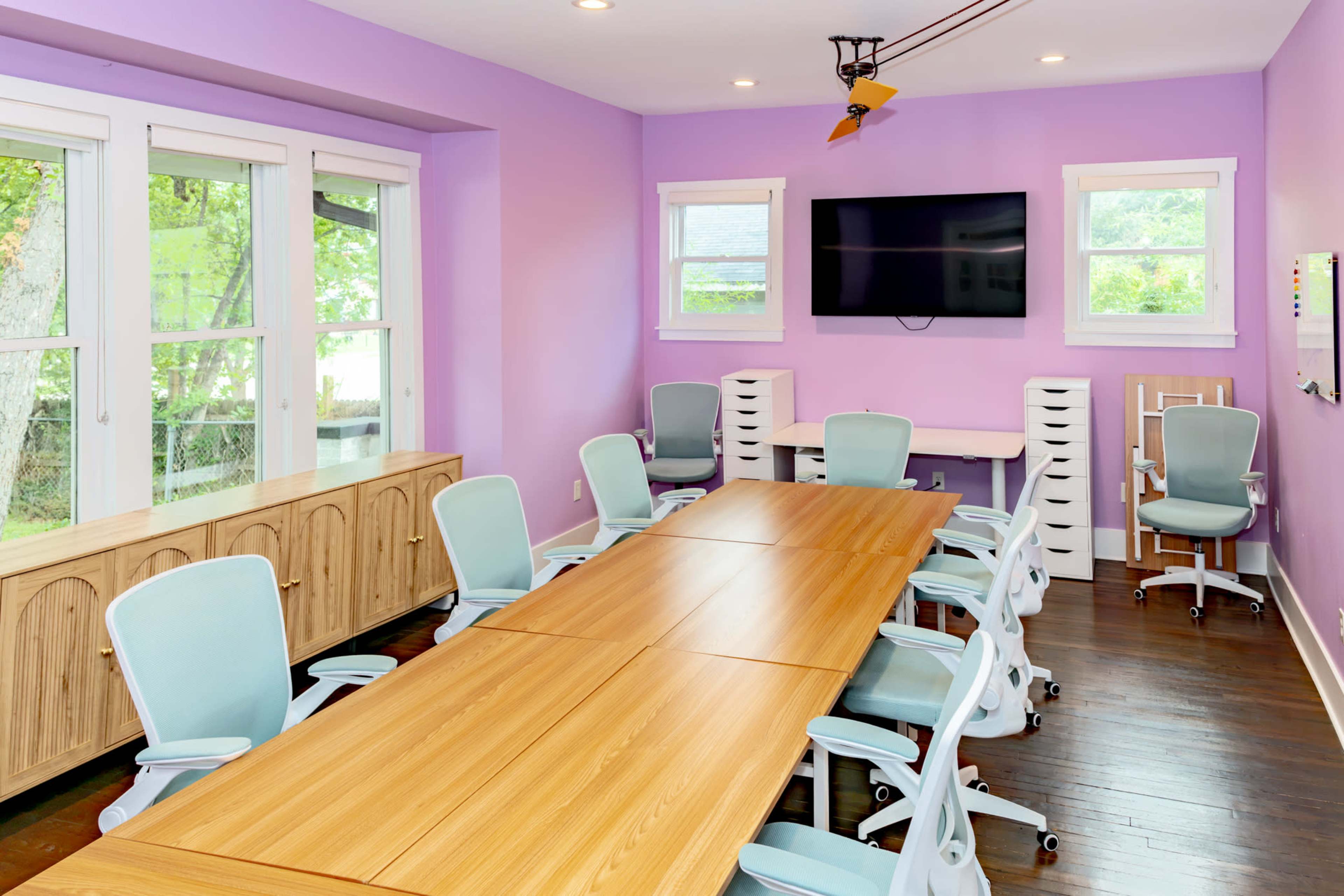 The image shows a meeting room with a long wooden table surrounded by light blue office chairs, purple walls, and a wall-mounted television.