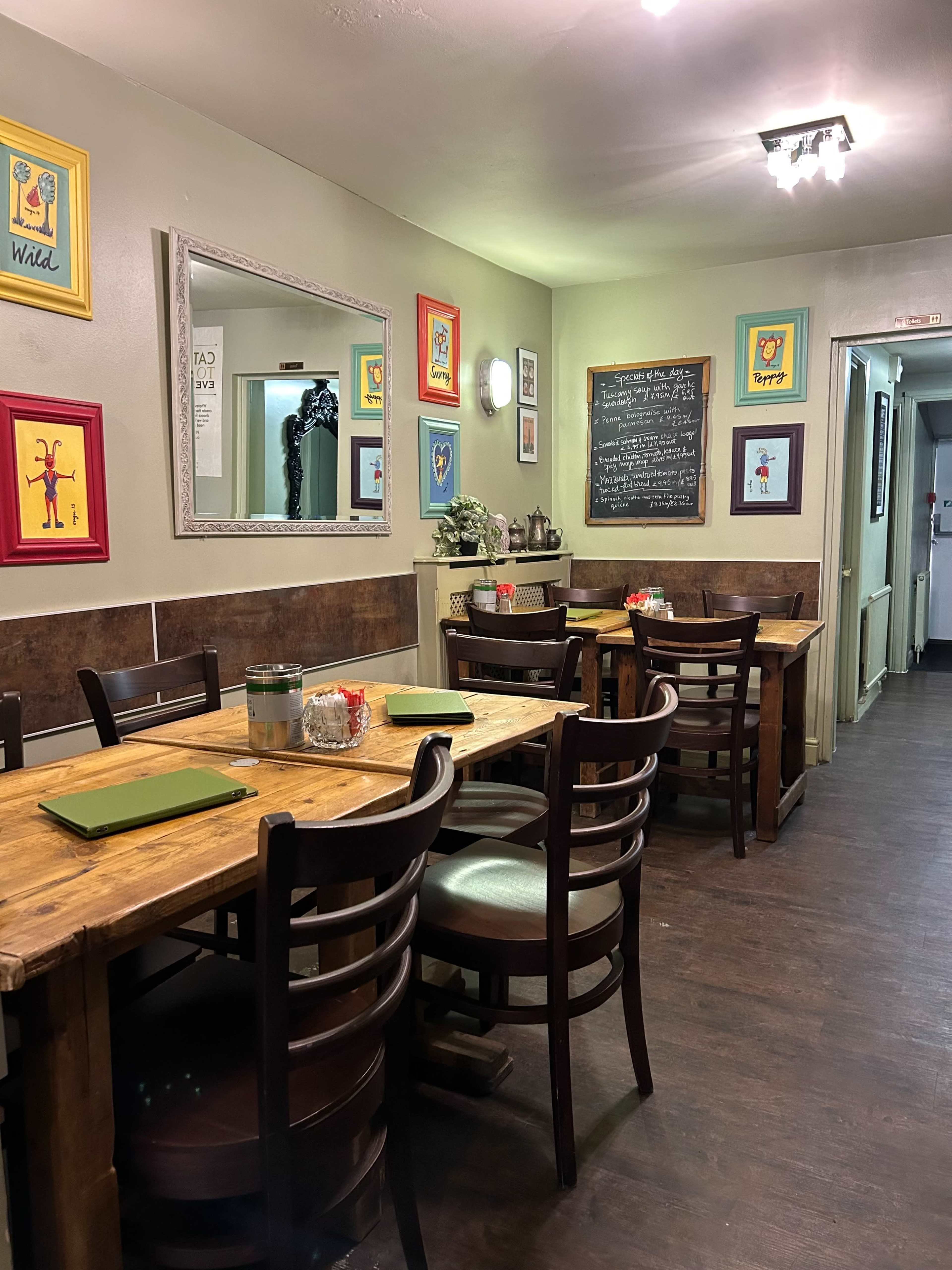 A rustic dining area of a restaurant with wooden tables and chairs, decorated walls featuring colorful framed art, and a chalkboard menu.