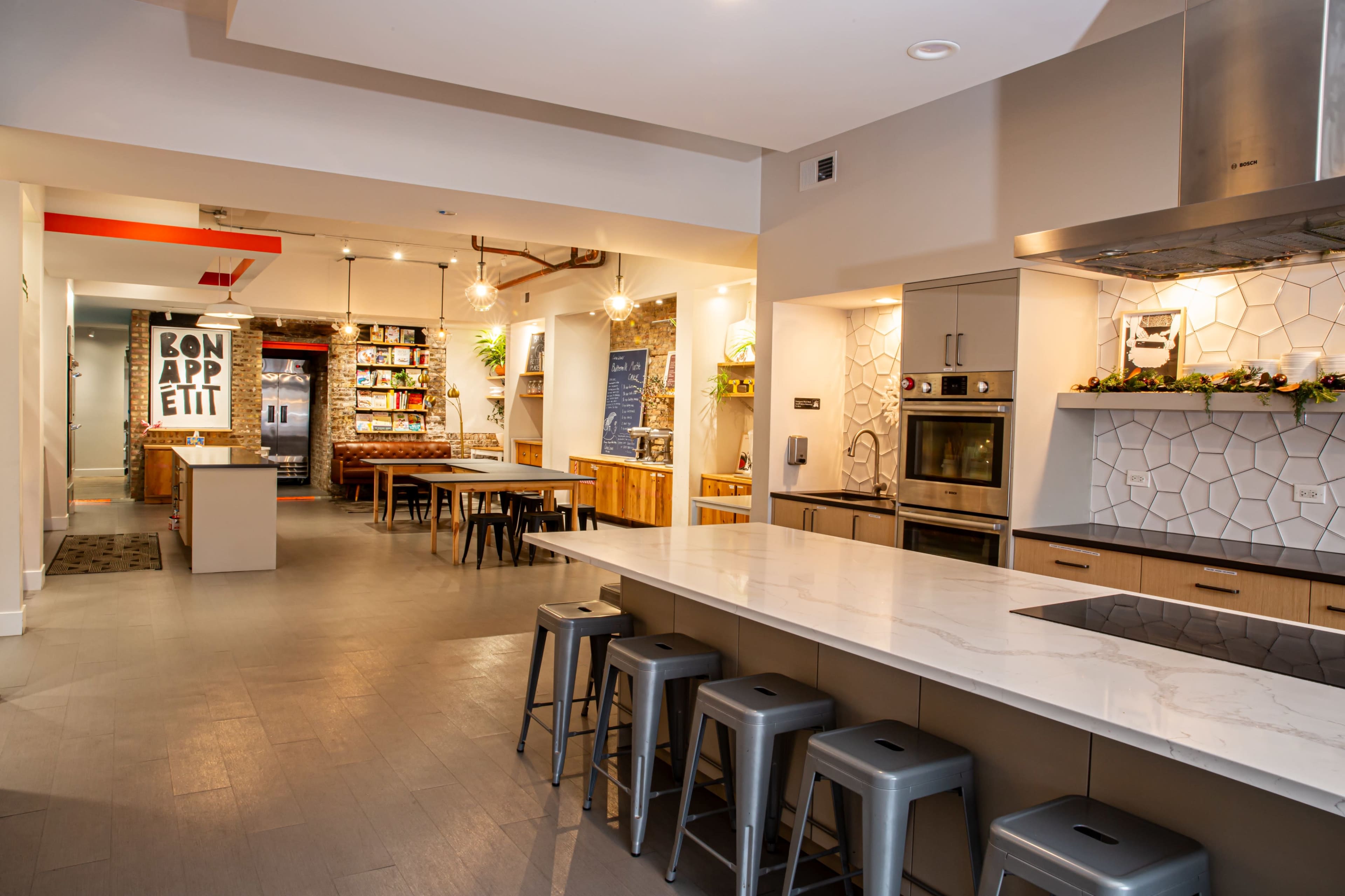 A modern kitchen and dining area with a large island, steel stools, and wood accents, leading to a room with shelving and a chalkboard wall.