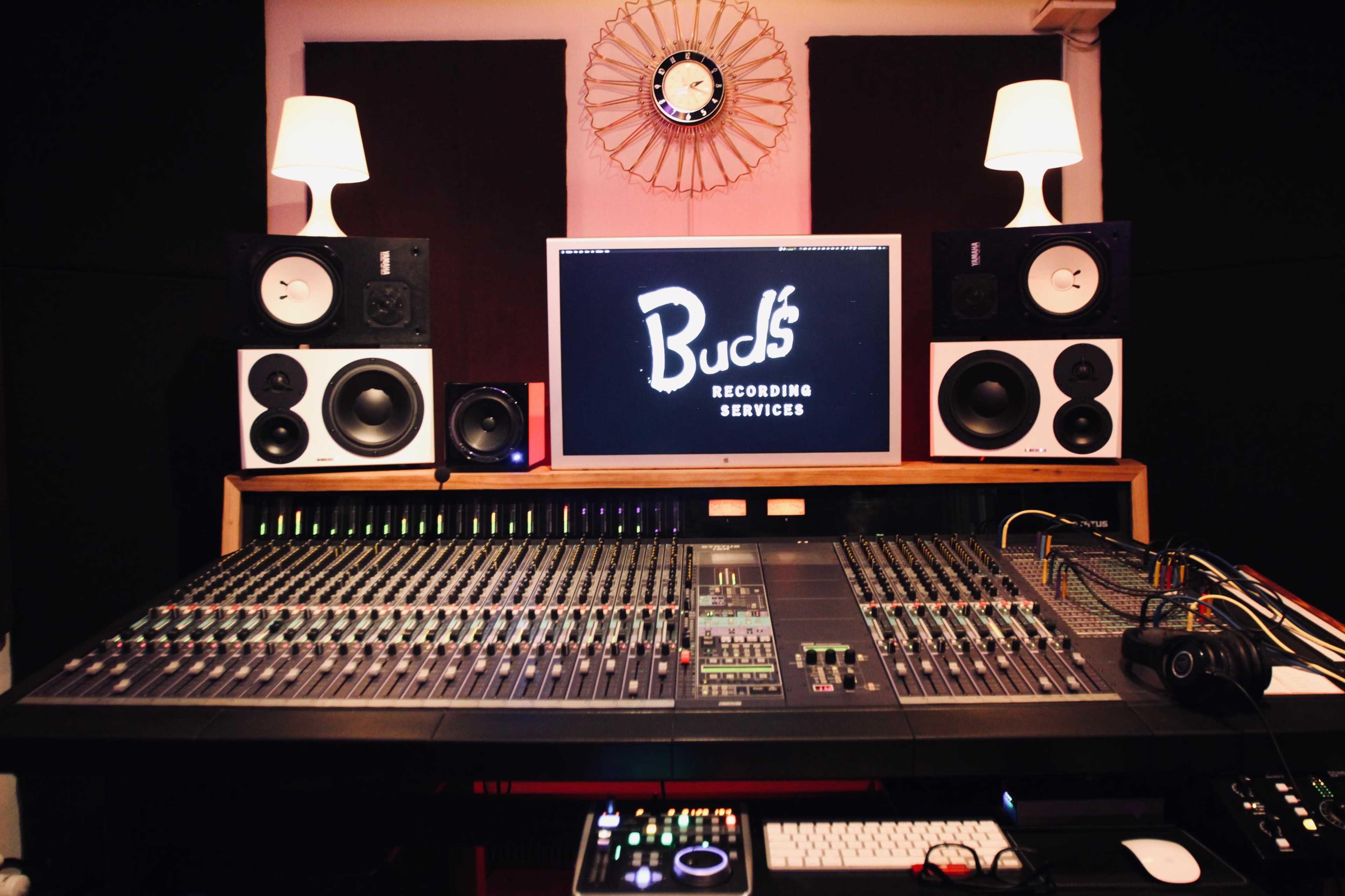 The image shows a recording studio with a large mixing console, a computer monitor displaying "Bud's Recording Services," speakers on either side, and various audio equipment.