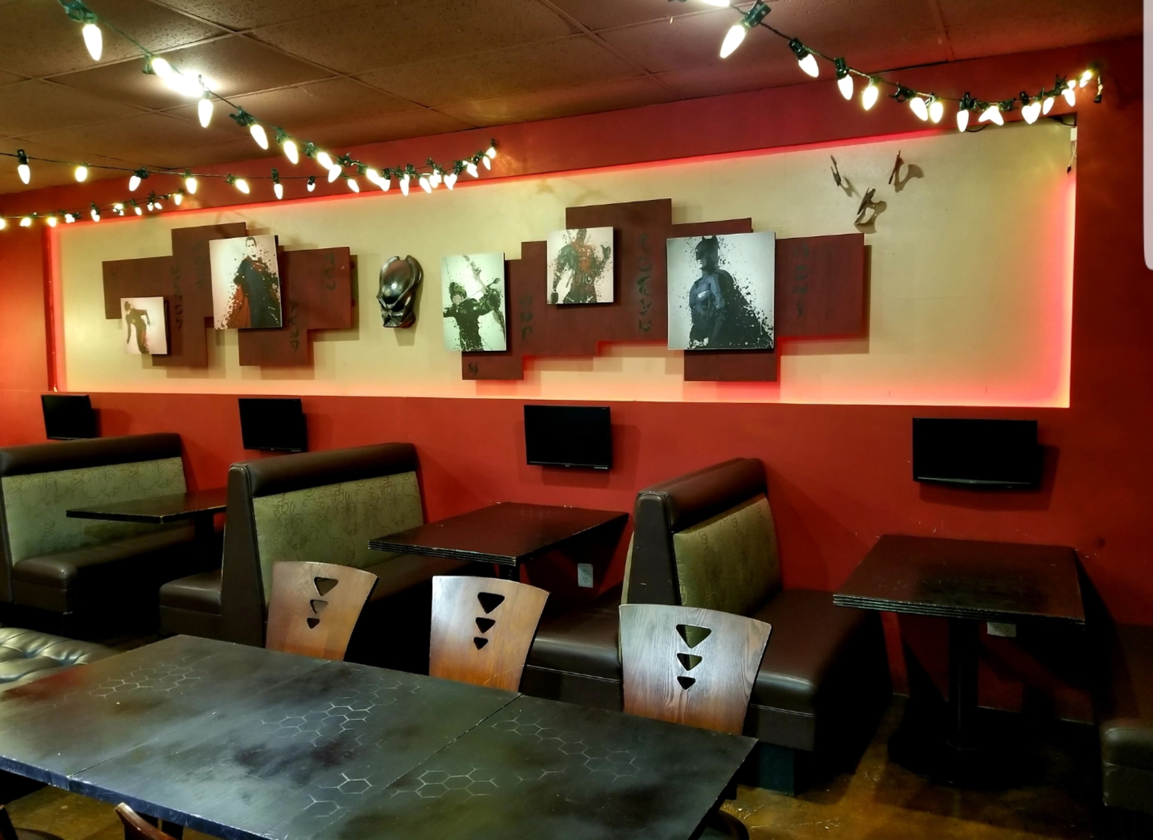 The image shows a café interior with dark wood tables, green upholstered booths, and framed artworks mounted on a red wall, illuminated by string lights.