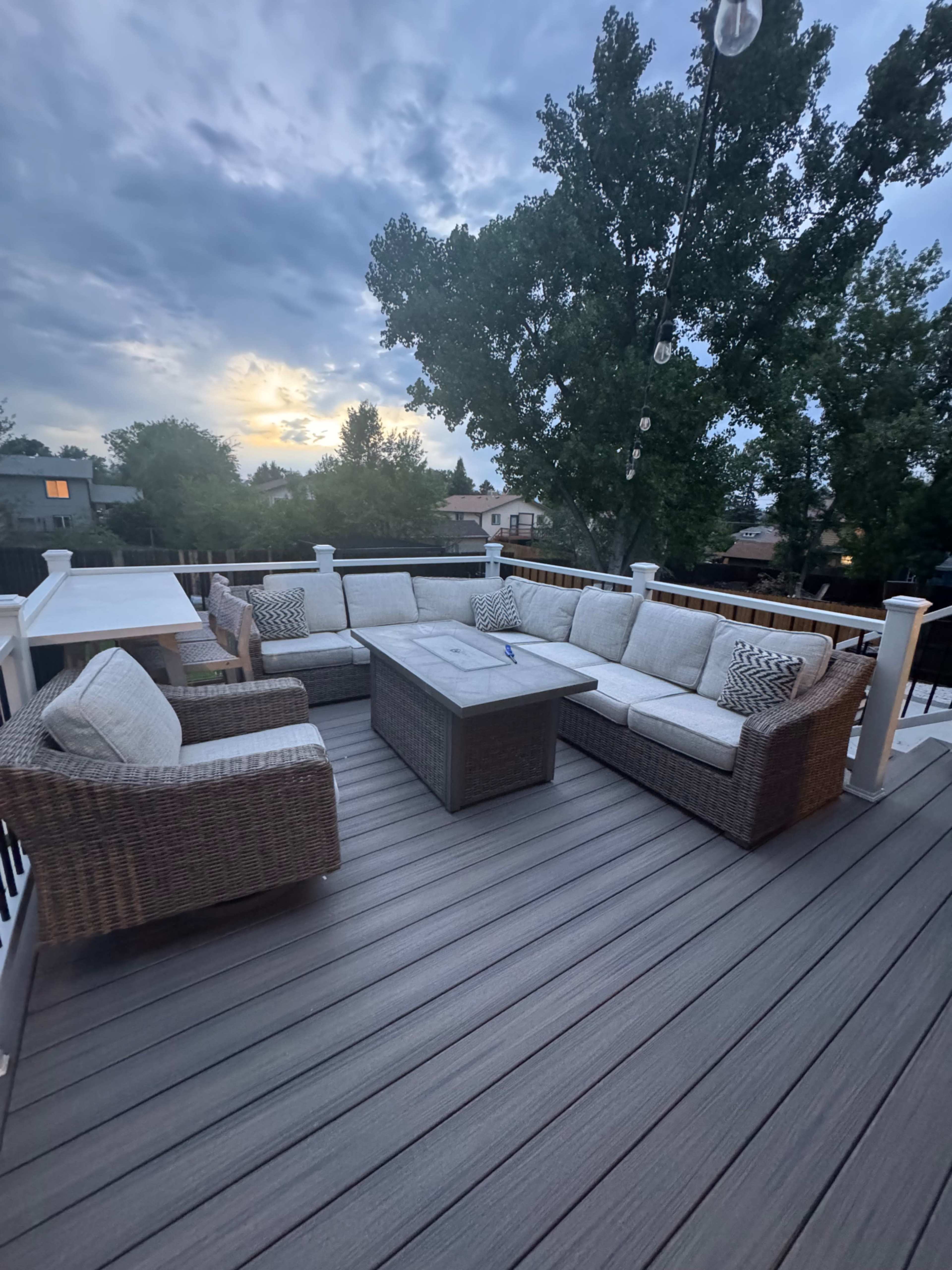 The image shows a cozy outdoor seating area with a large sectional sofa, a coffee table, and a view of trees and a cloudy sky.