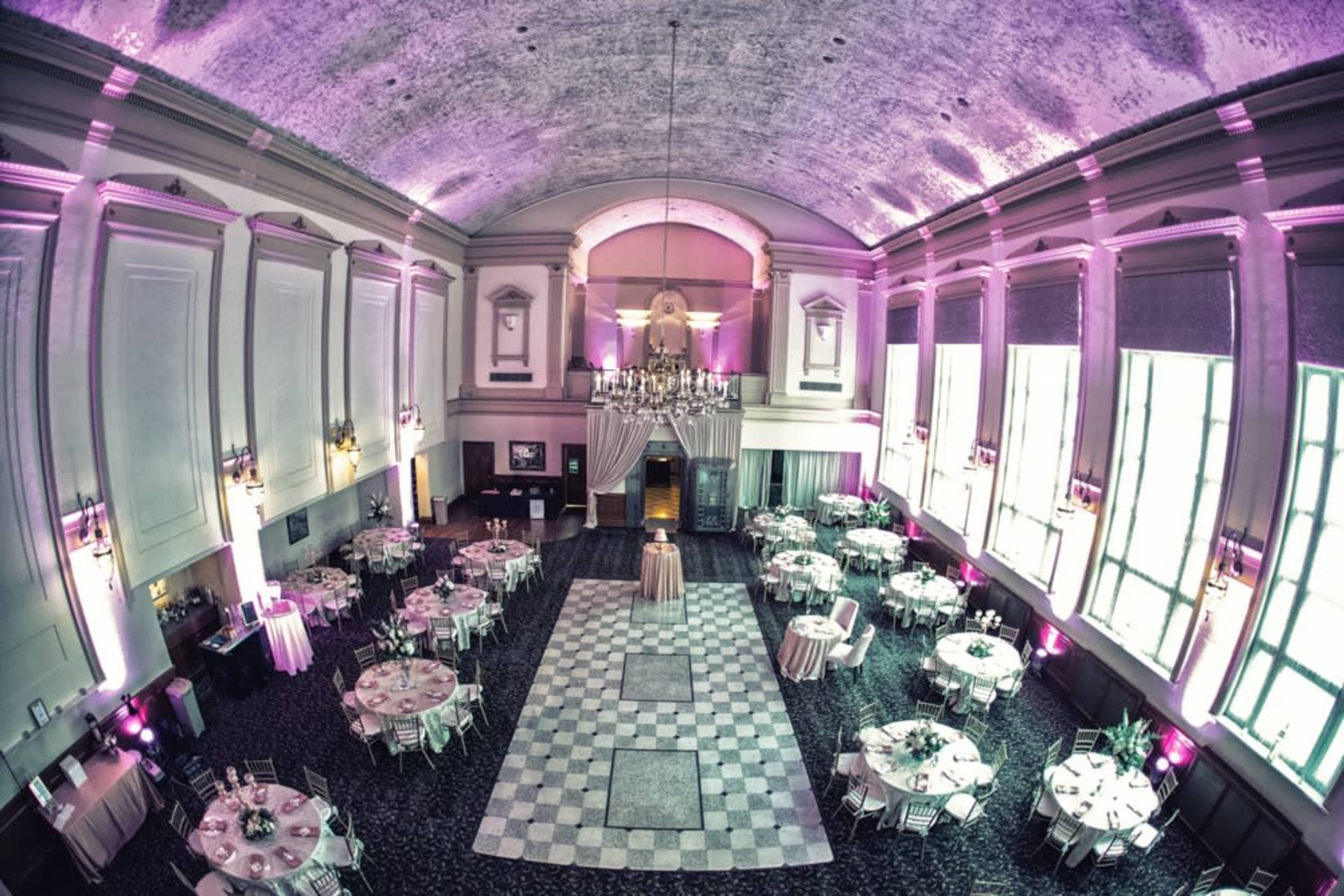 An elegantly arranged banquet hall with round tables, a checkered dance floor, and purple lighting illuminating the high ceiling.