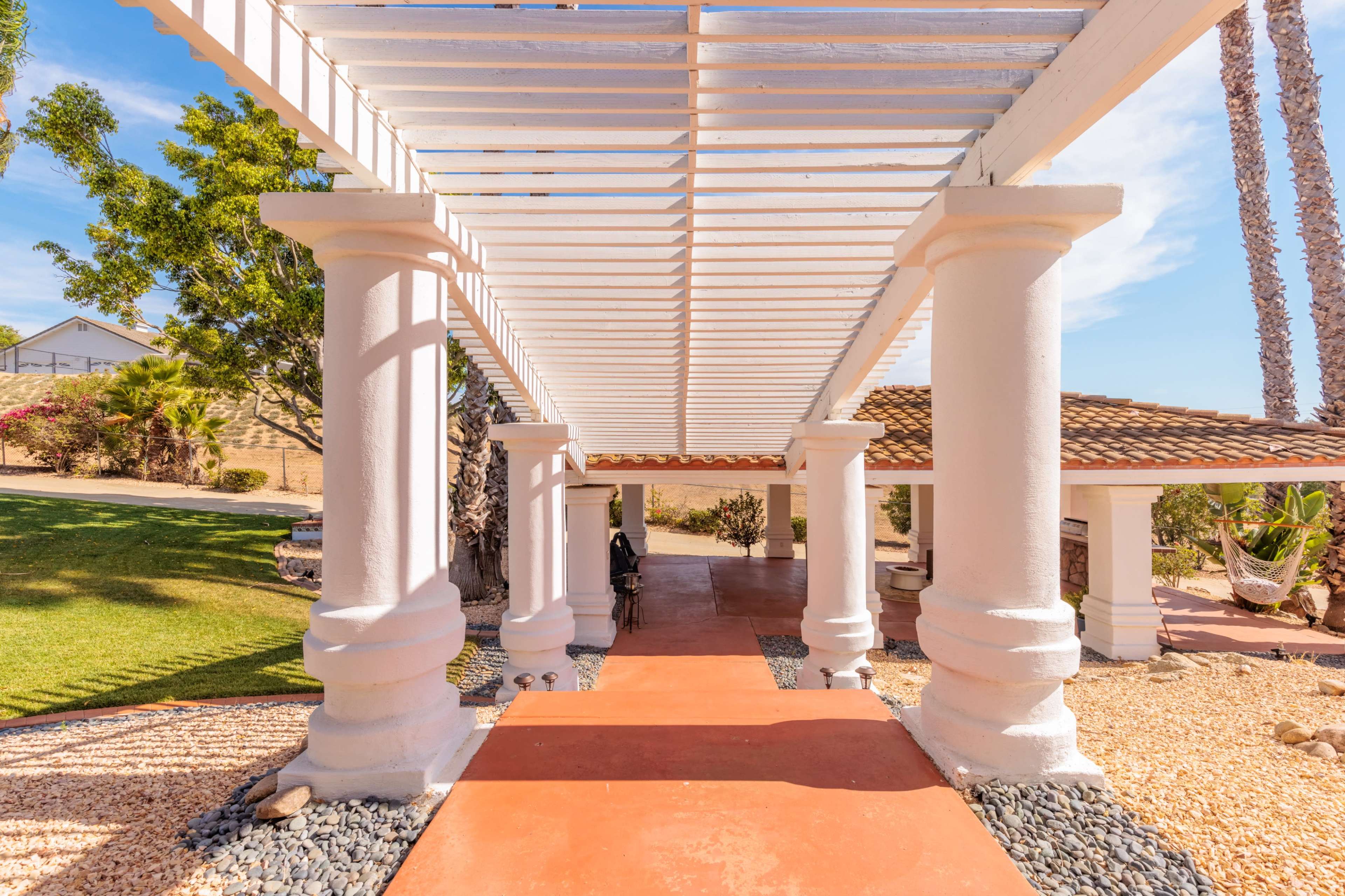 A walkway supported by white columns, leading through a pergola with open slats and surrounded by landscaped gardens.