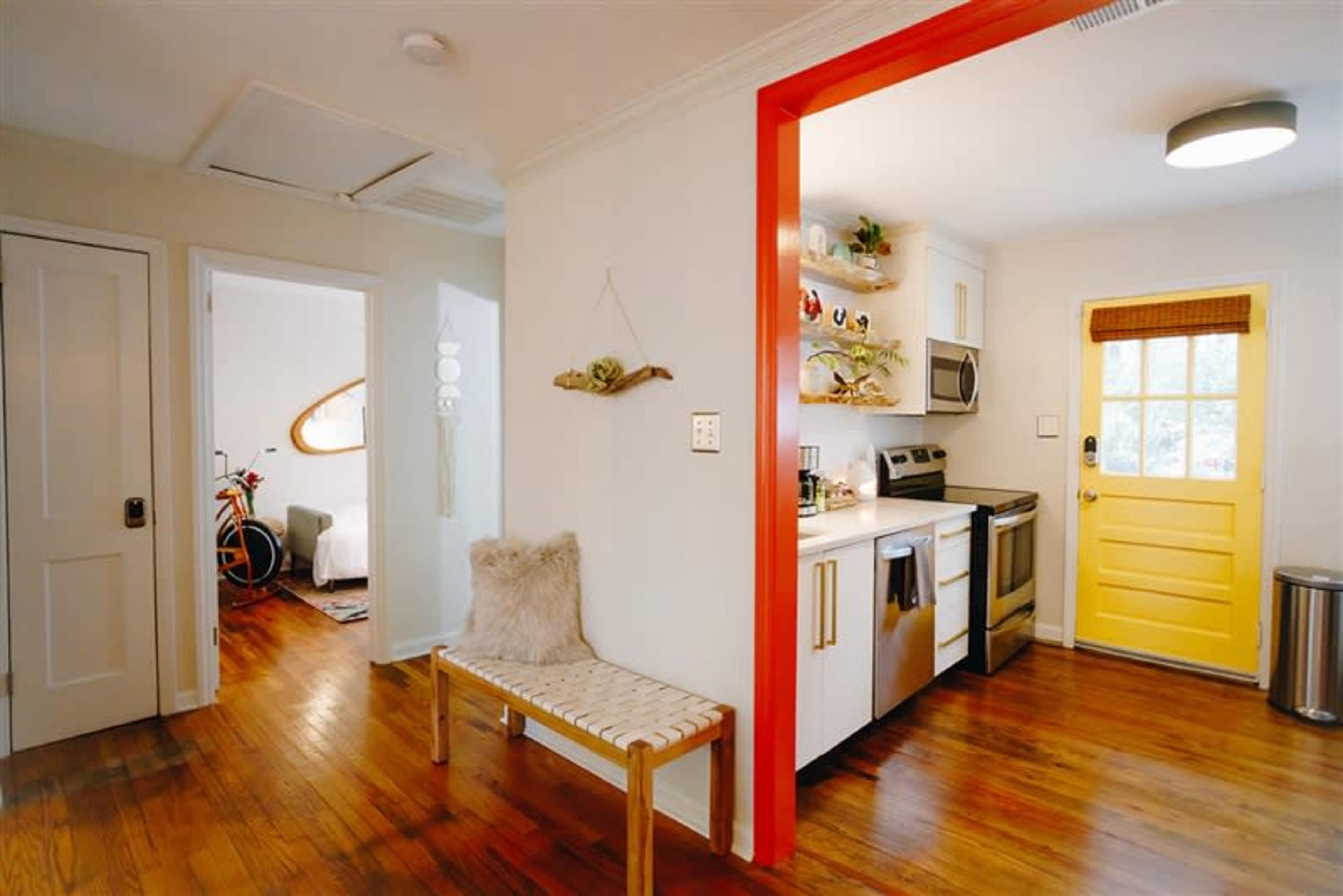 A bright kitchen with stainless steel appliances, a yellow door, and a wooden bench in a hallway leading to another room.