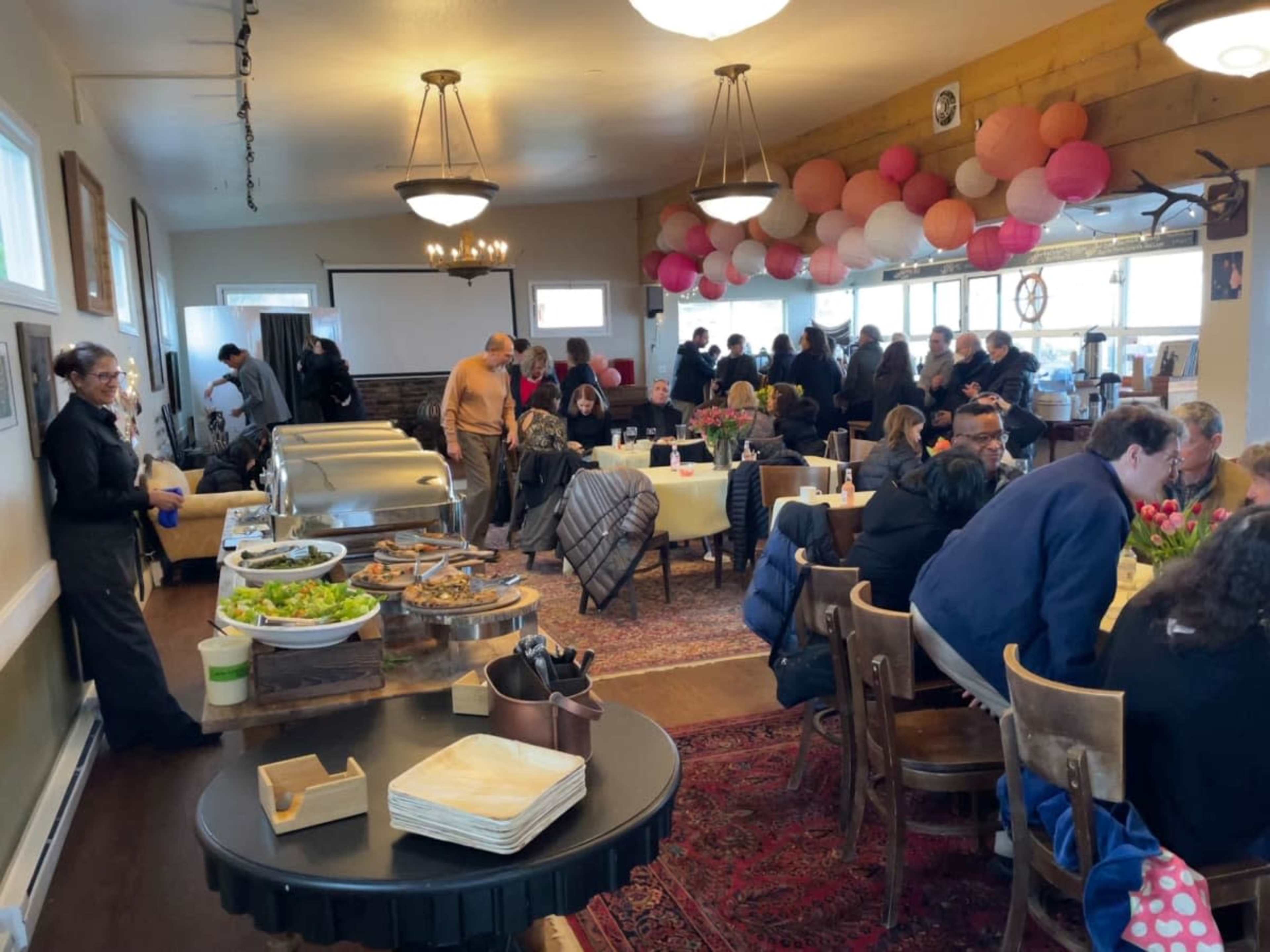 A crowd of people gathers in a warmly lit dining room filled with tables, enjoying a buffet-style meal and decorated with pink and white paper lanterns.