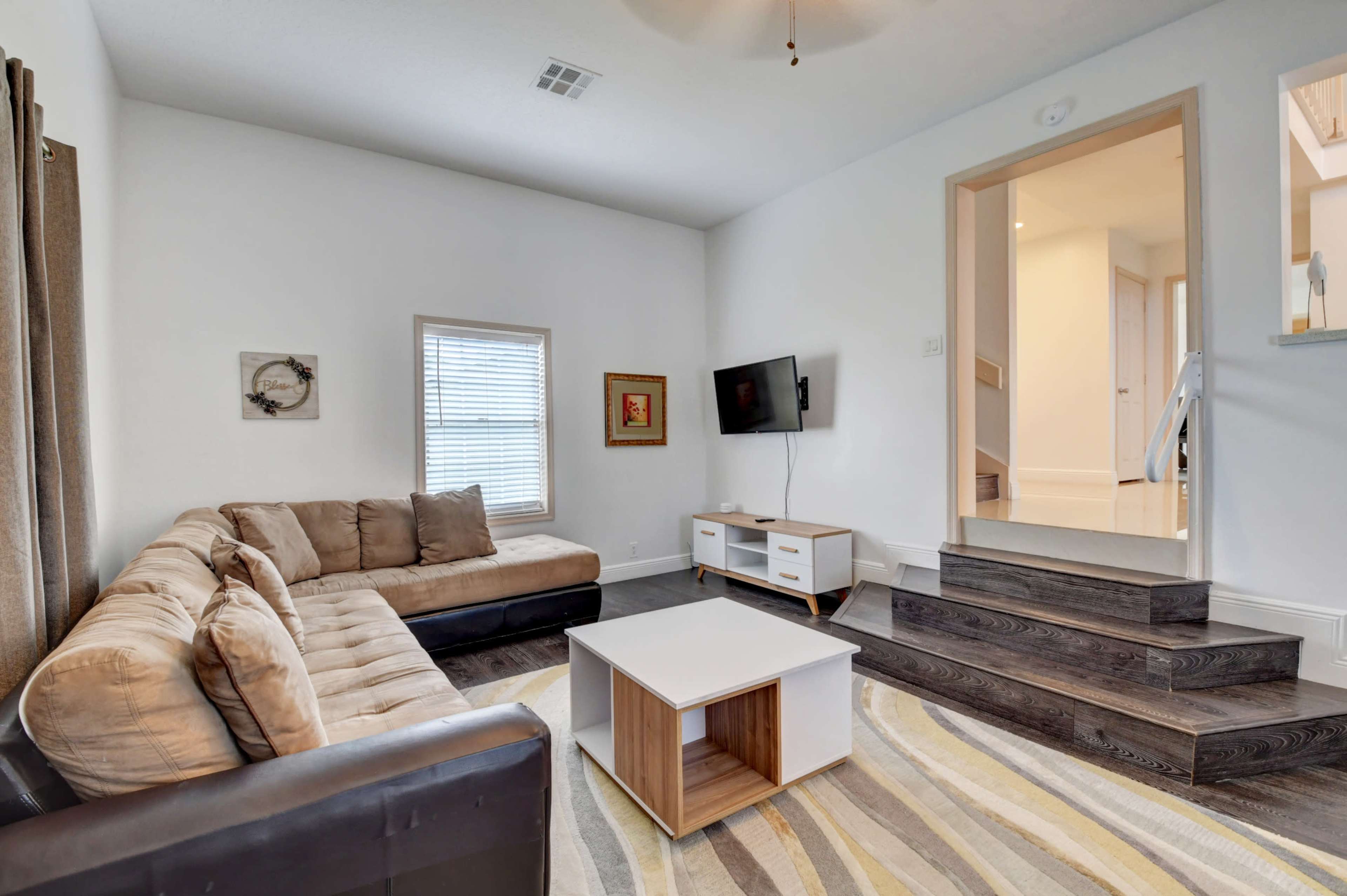 A living room features a sectional sofa, a coffee table, and a mounted television, with stairs leading to another room.