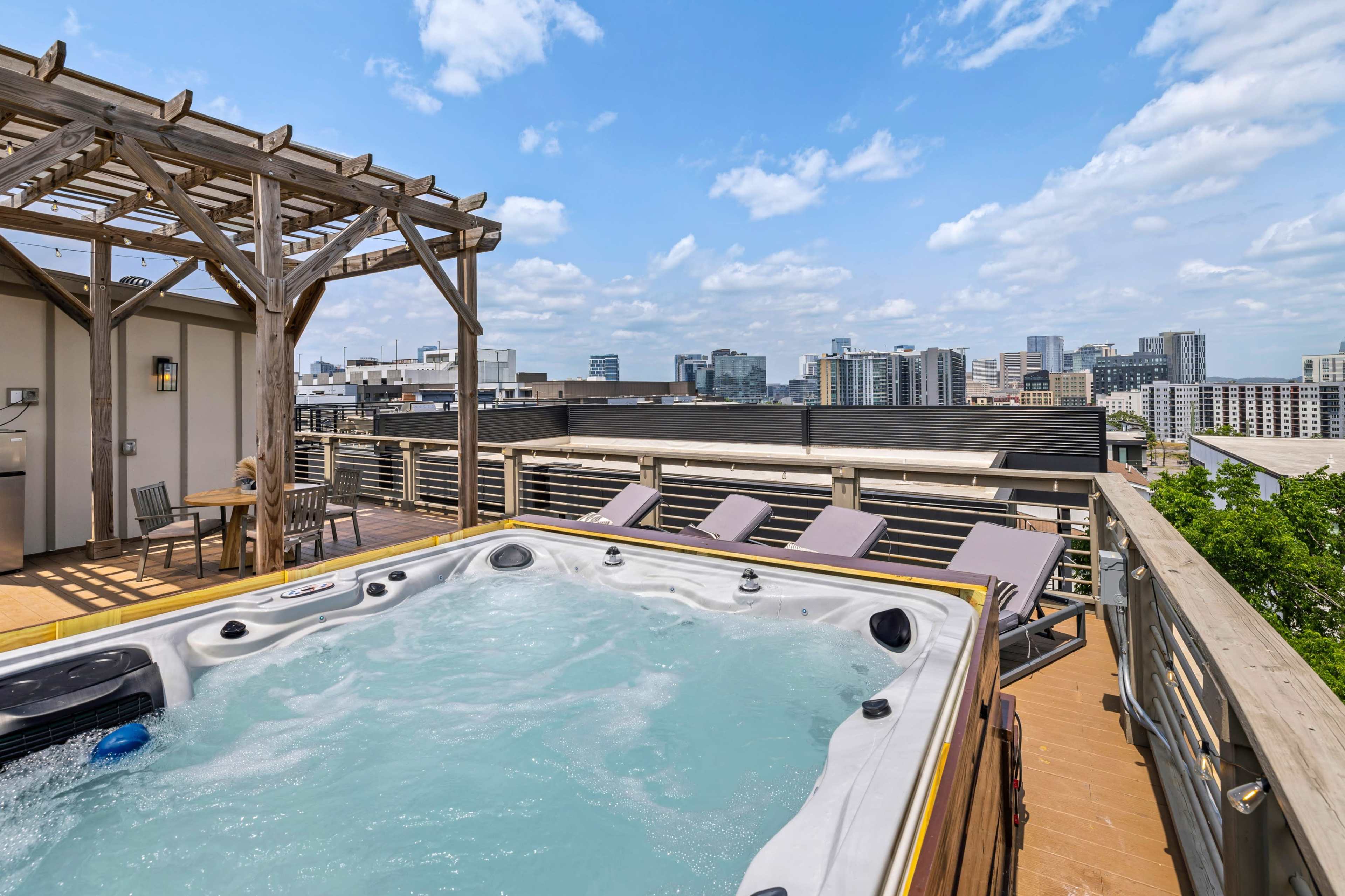 A rooftop deck features a hot tub with city skyline views and lounge chairs under a pergola.