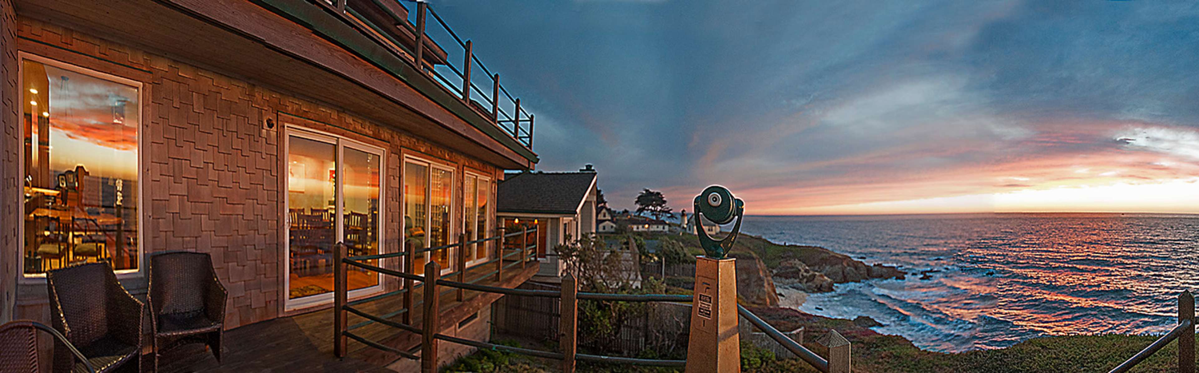 A coastal view from a wooden terrace with chairs overlooking the ocean at sunset, featuring a telescope mounted on a post.