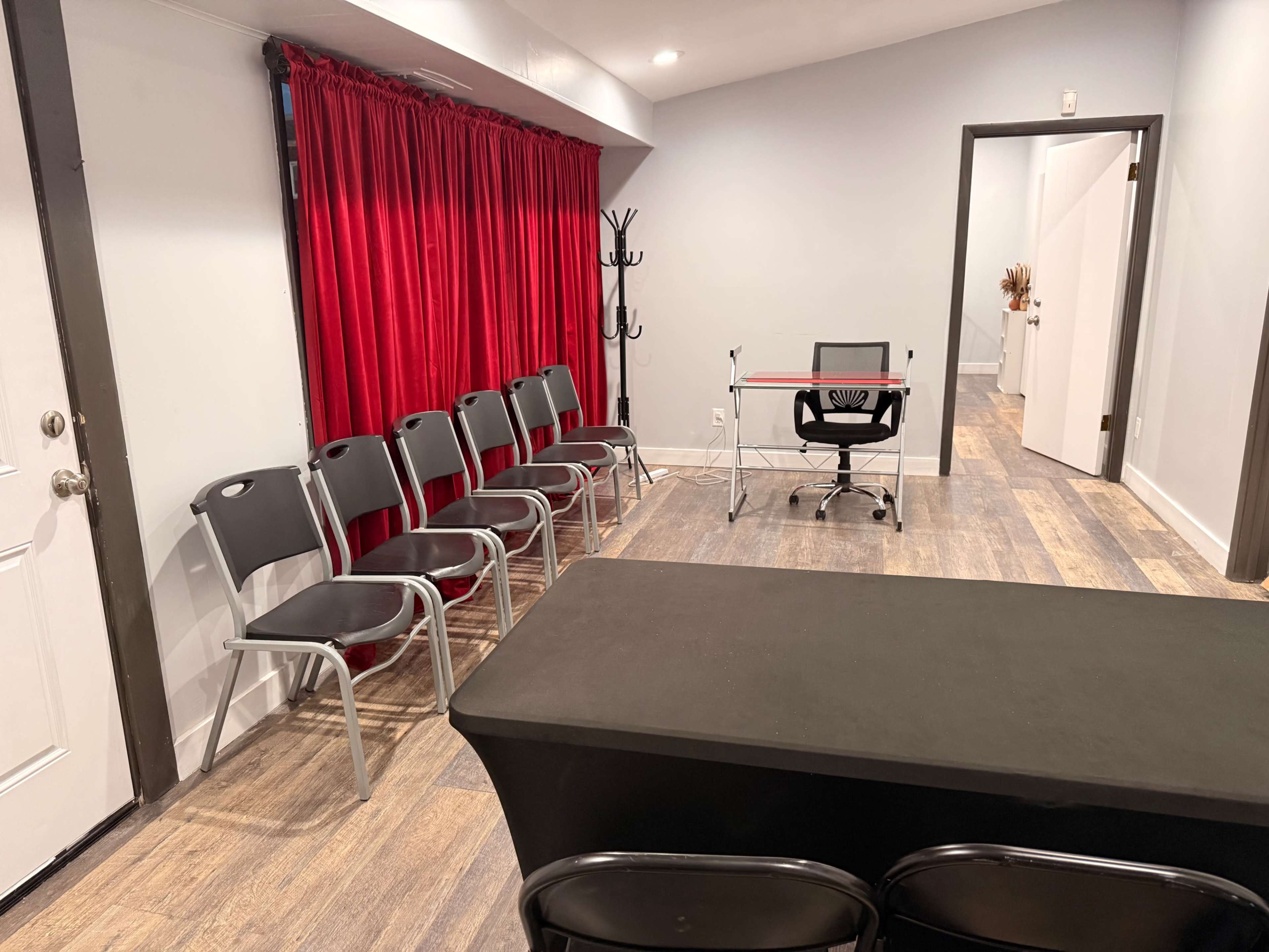 The image shows an interior room with a row of metal chairs, a black table, a desk with a chair, and a red curtain along one wall.