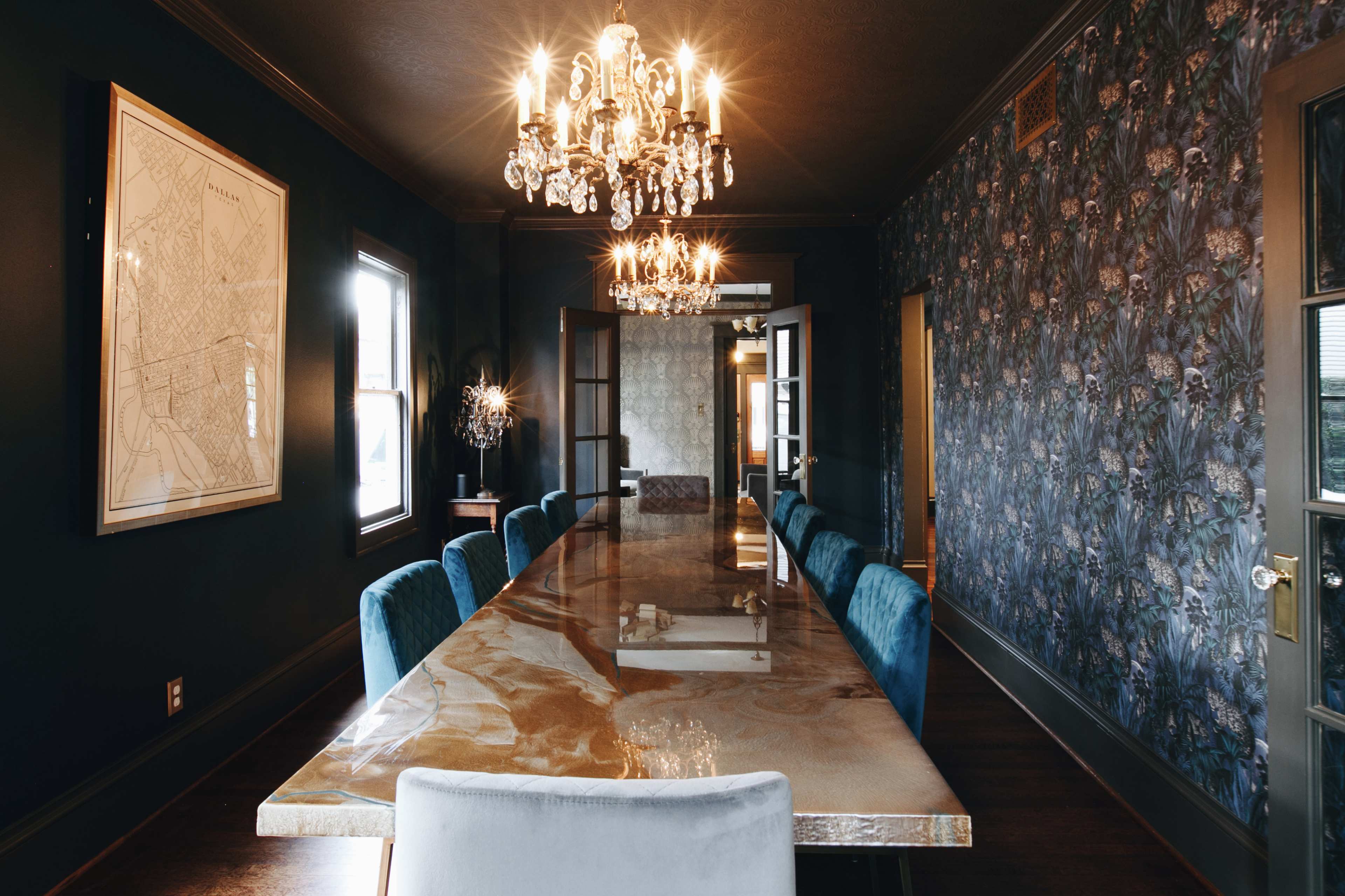 A long table with a glossy surface and blue chairs is centrally positioned in a dimly lit dining room adorned with floral wallpaper and chandeliers.