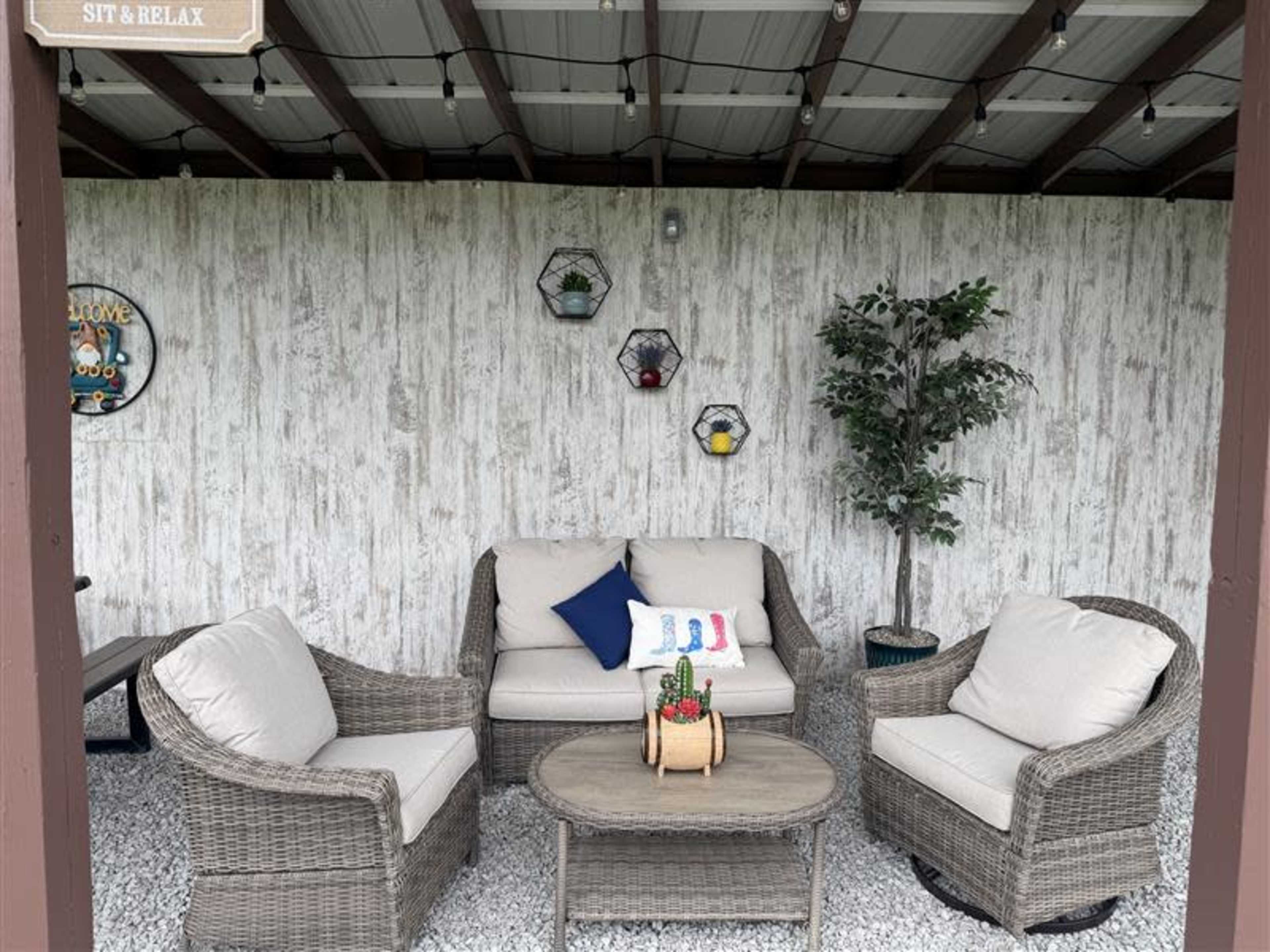 A seating area with a round table and four wicker chairs is arranged under a covered patio featuring decorative plants and wall hangings.
