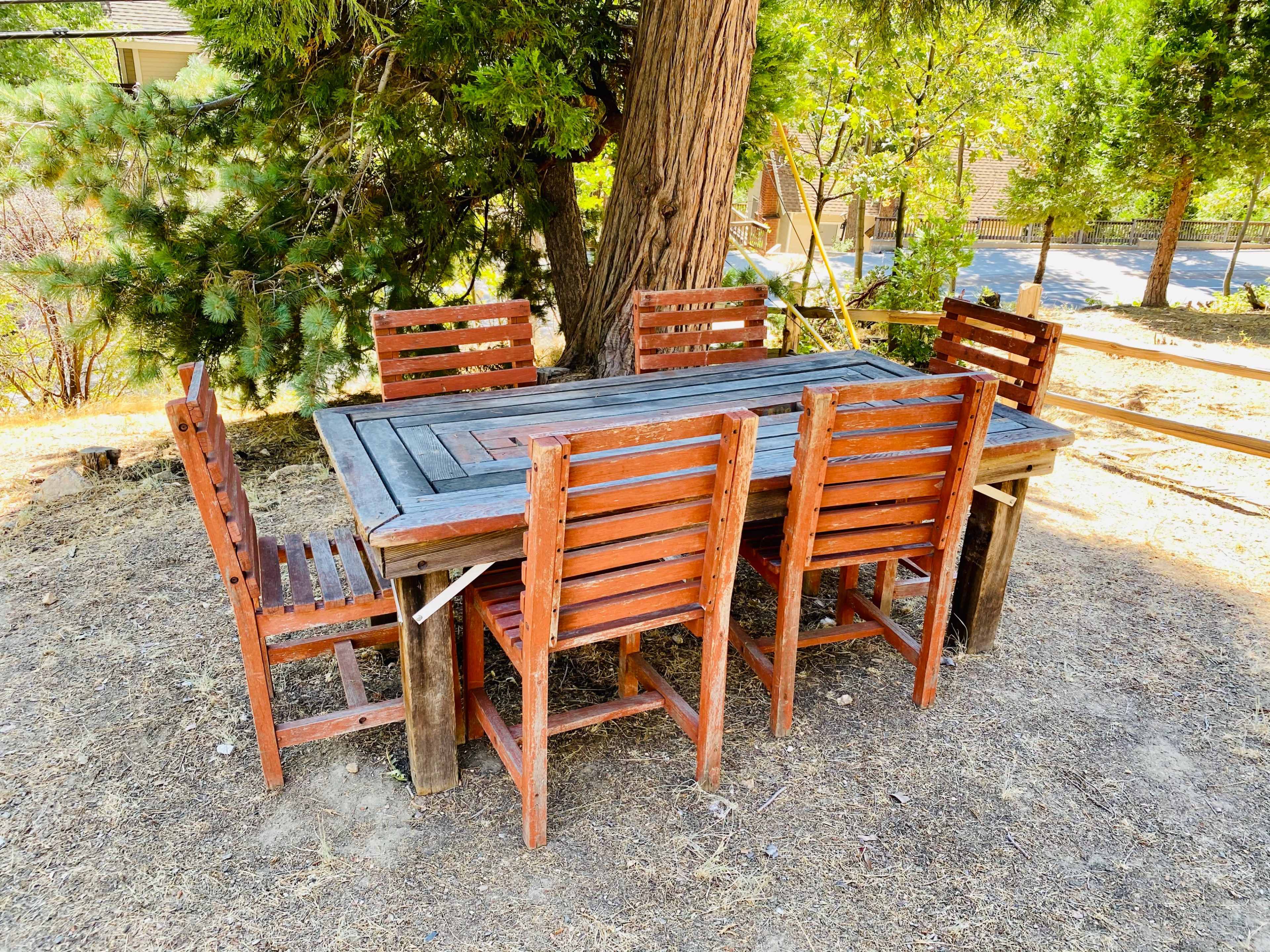 A wooden outdoor dining table surrounded by six matching chairs sits on bare ground under a tree.