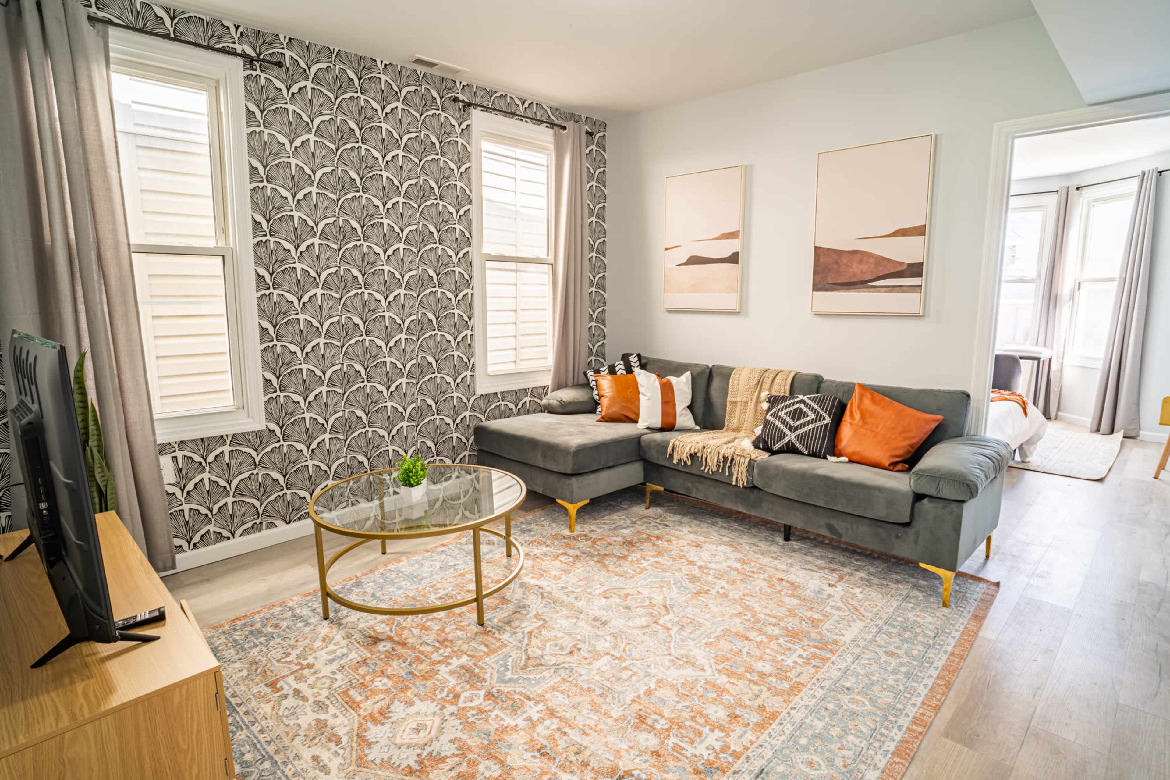 A modern living room featuring a gray sectional sofa, a round glass coffee table, decorative artwork on the walls, and large windows allowing natural light.