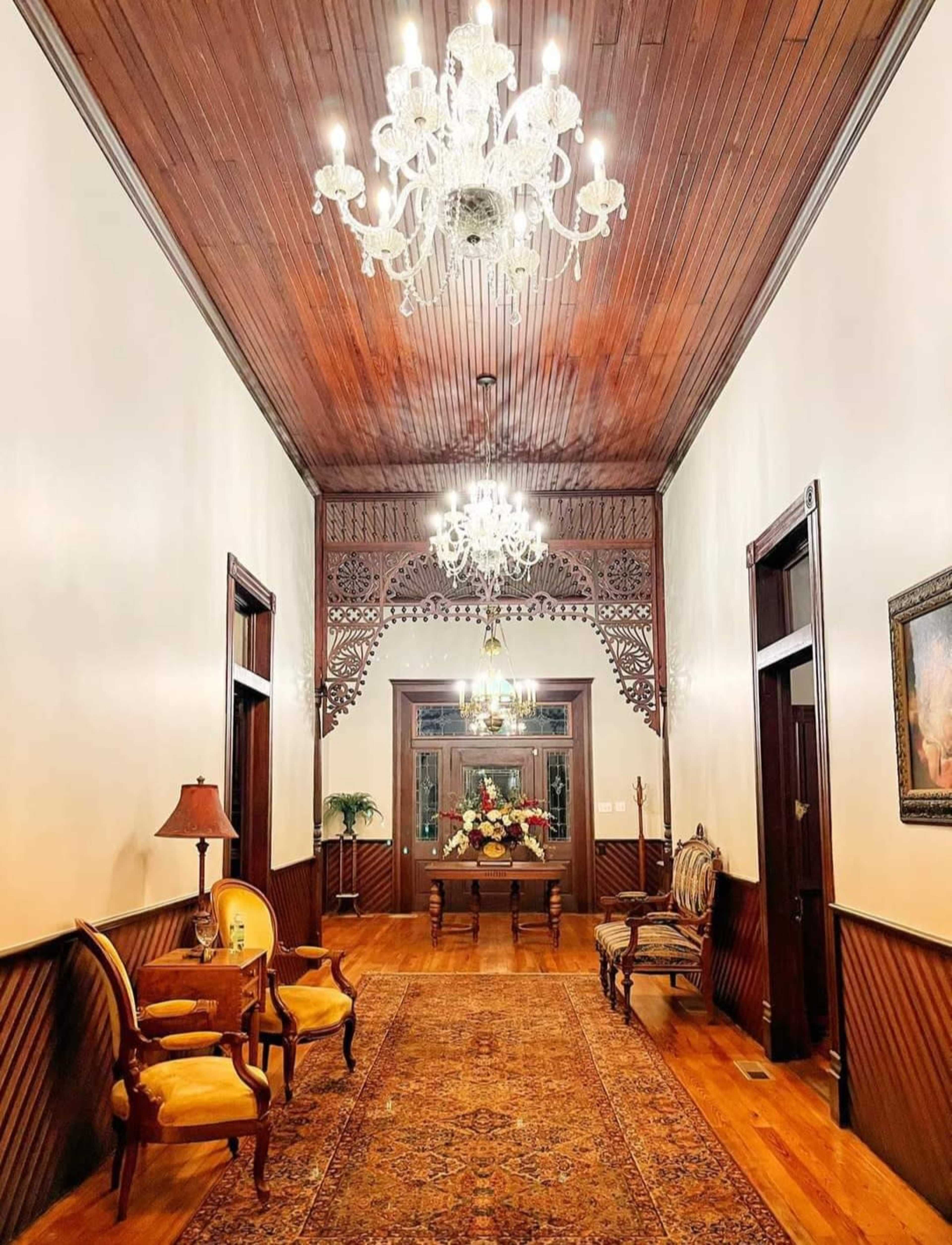 A long hallway featuring wooden paneled walls, ornate chandeliers, and elegant furniture, with a decorative mirror at the far end.