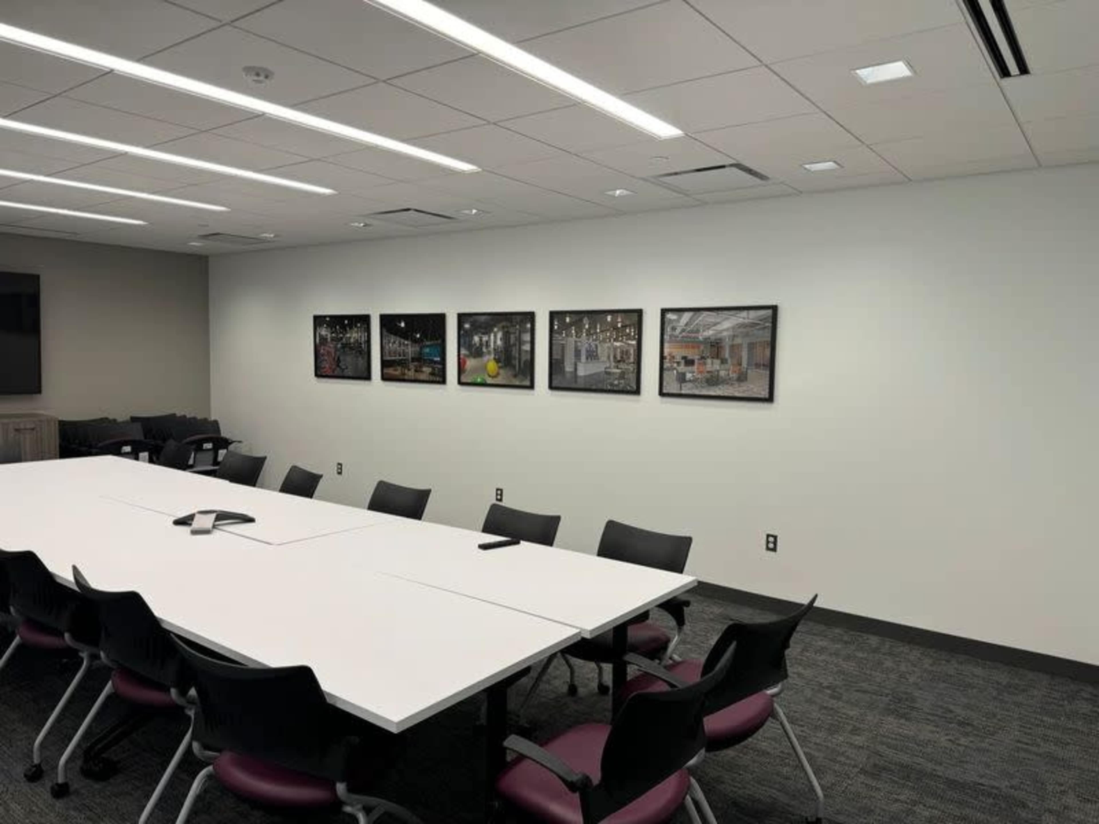 A modern conference room features a long white table surrounded by black chairs, with framed photographs hanging on the wall.
