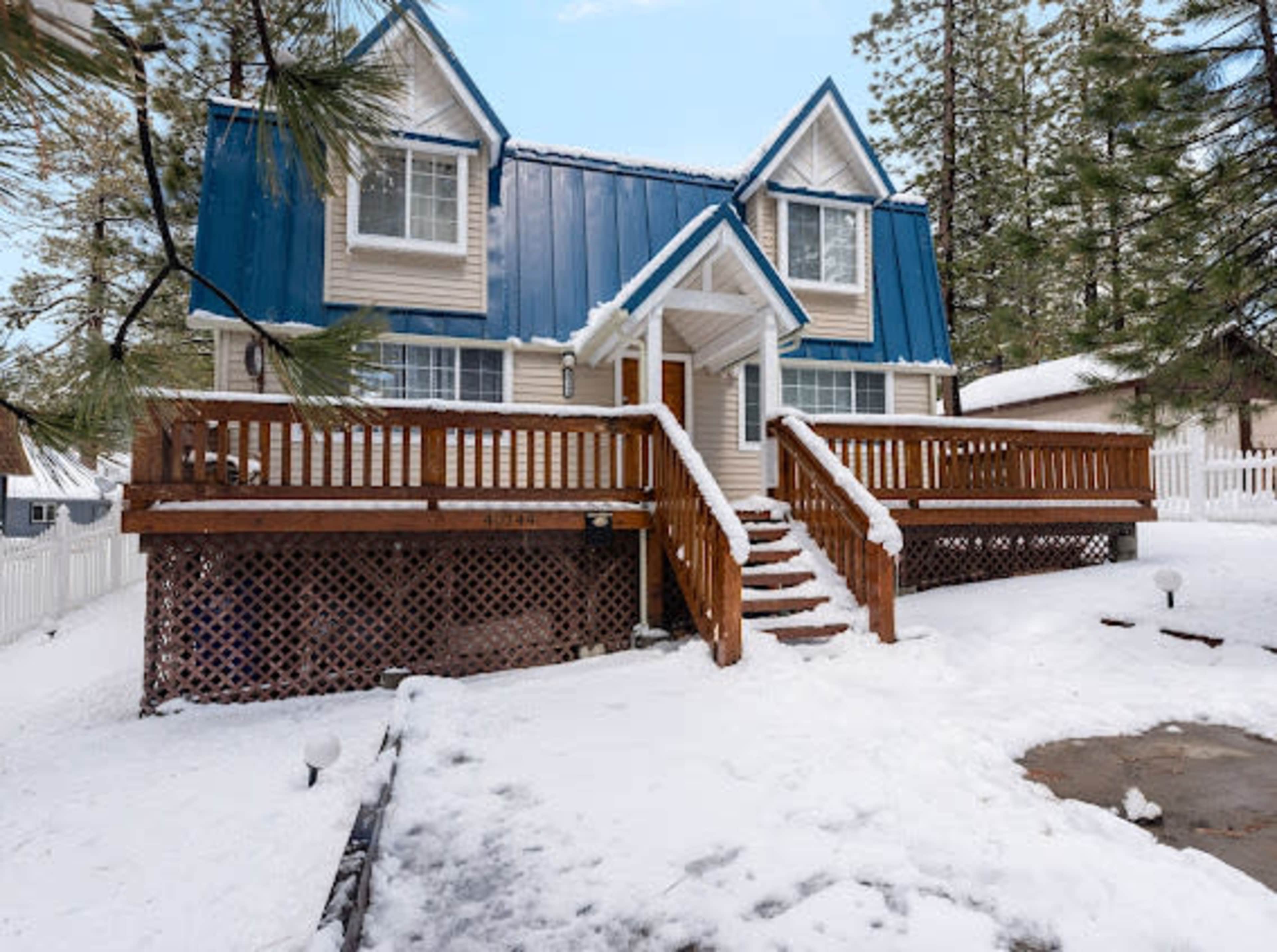 A two-story house with a blue metal roof and wooden deck is surrounded by snow and pine trees.