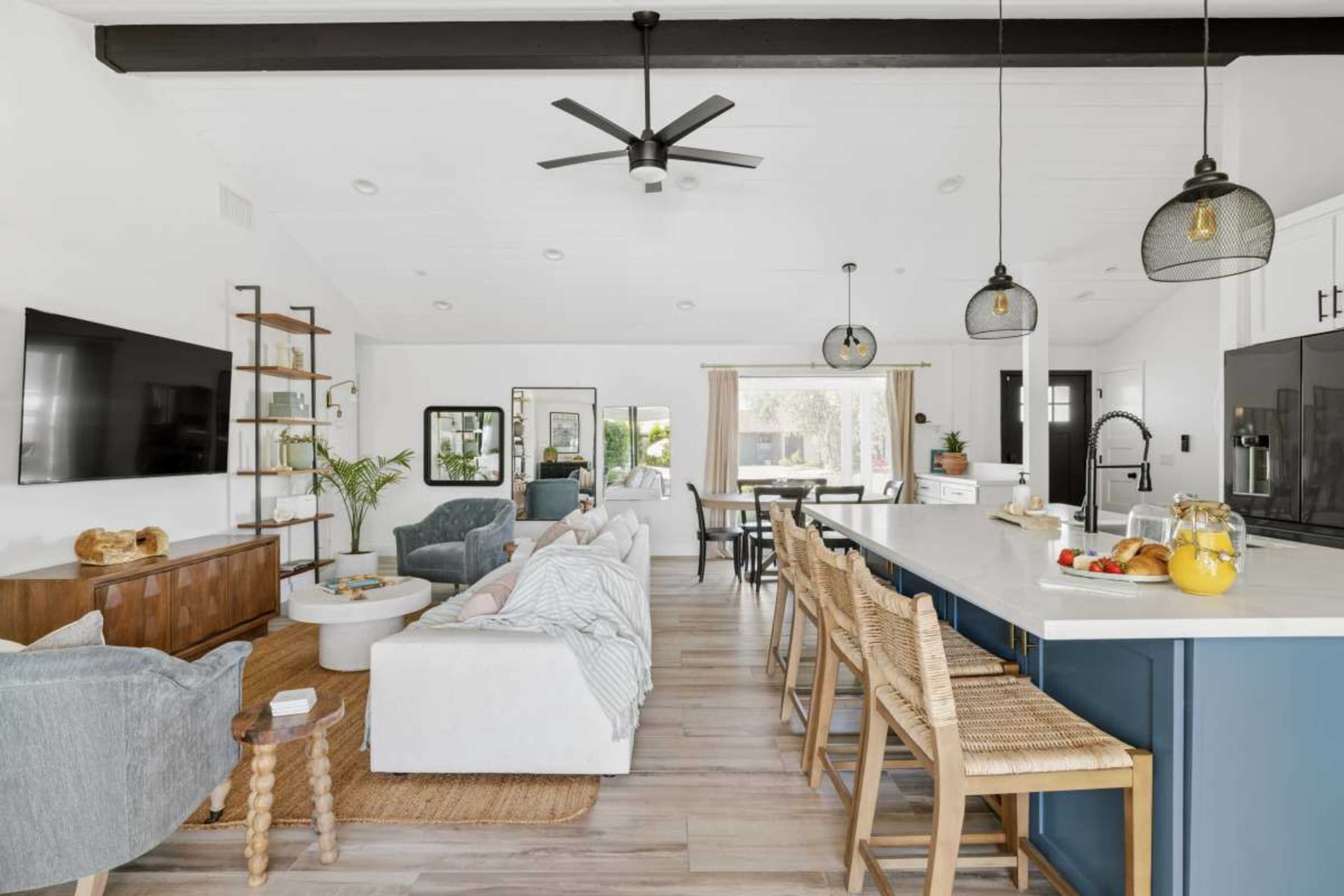 The image shows a modern, open-concept living space featuring a kitchen with bar seating, a dining area, and a cozy sitting area with light-colored furniture.