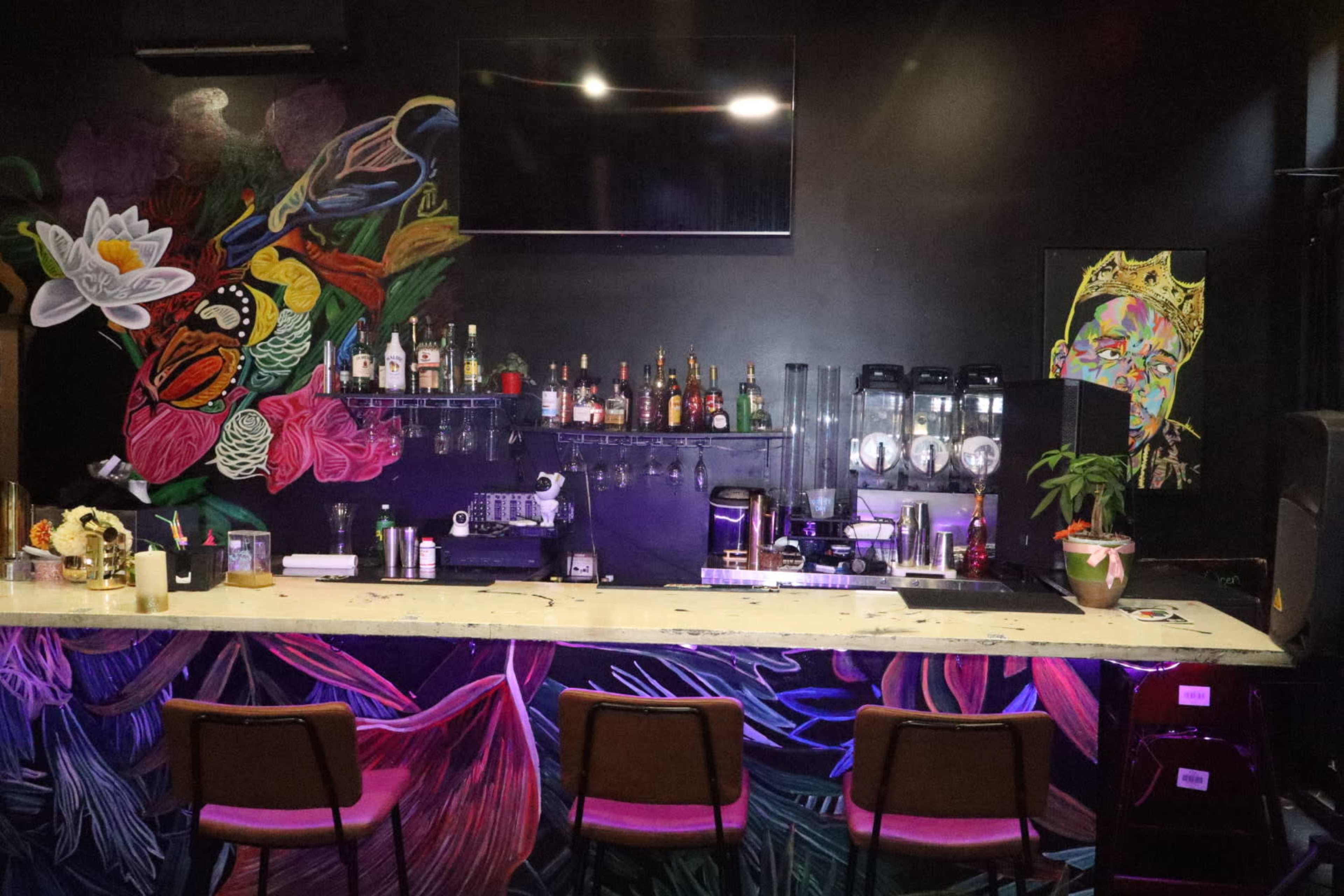 The image shows a bar area with a colorful mural on the wall, a selection of bottles displayed on shelves, and a few bar stools arranged in front of the counter.