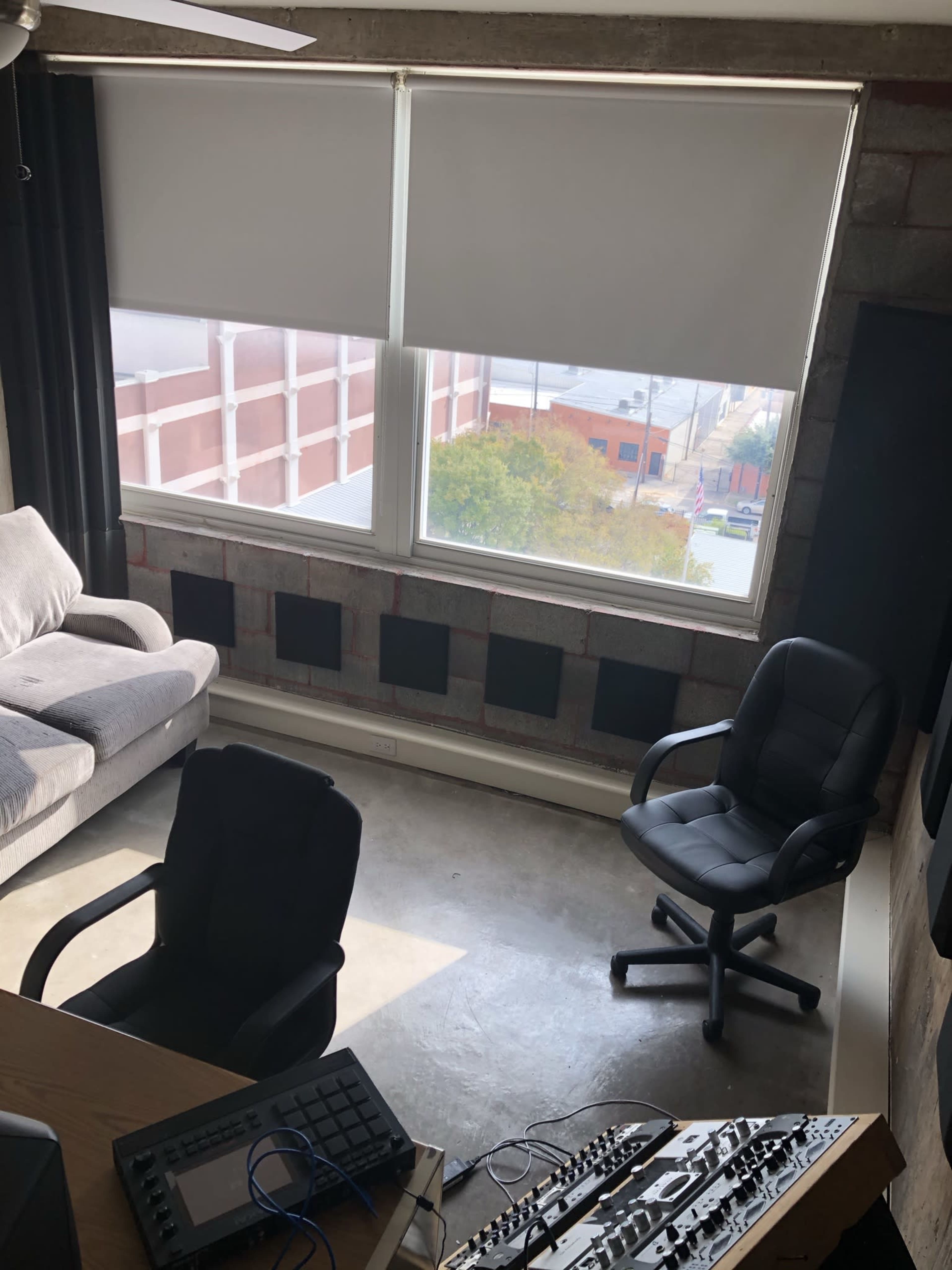 A spacious room with a gray couch, a black chair, and a window overlooking an exterior view, featuring muted light and concrete walls.