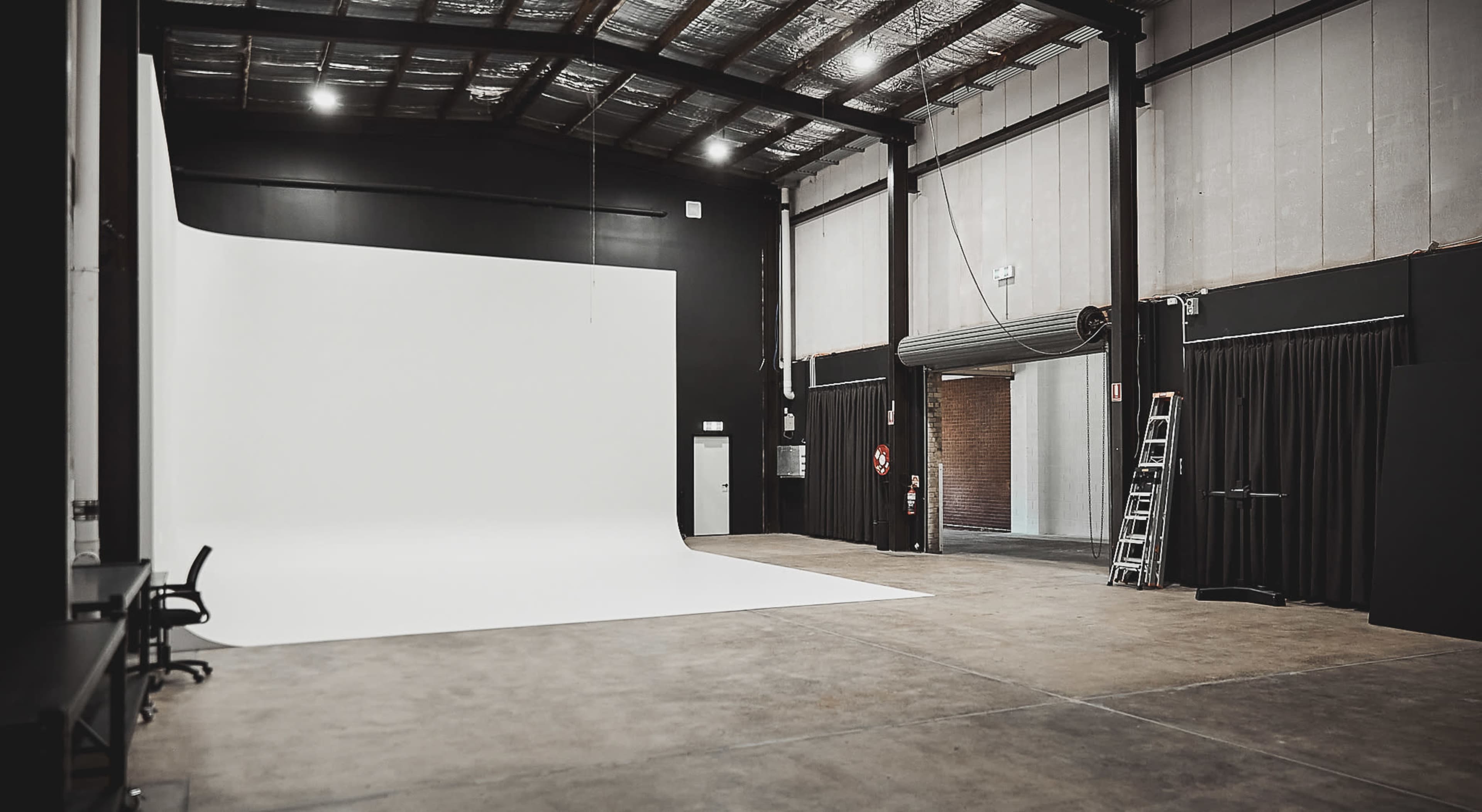 A spacious photography studio featuring a large white backdrop, black walls, and industrial lighting fixtures.