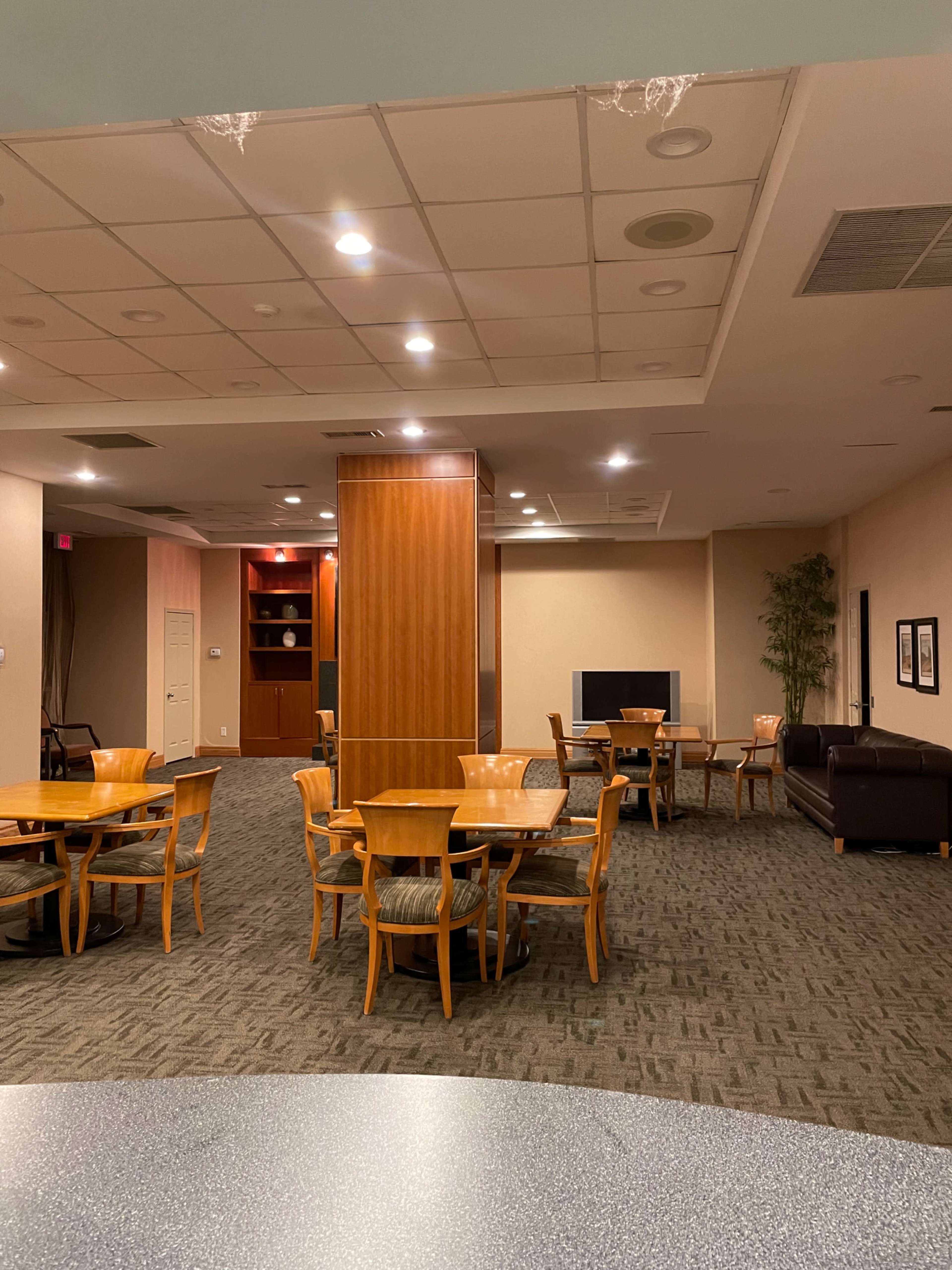 A spacious room with wooden tables and chairs, a large wooden column, a seating area, and shelves in the background.