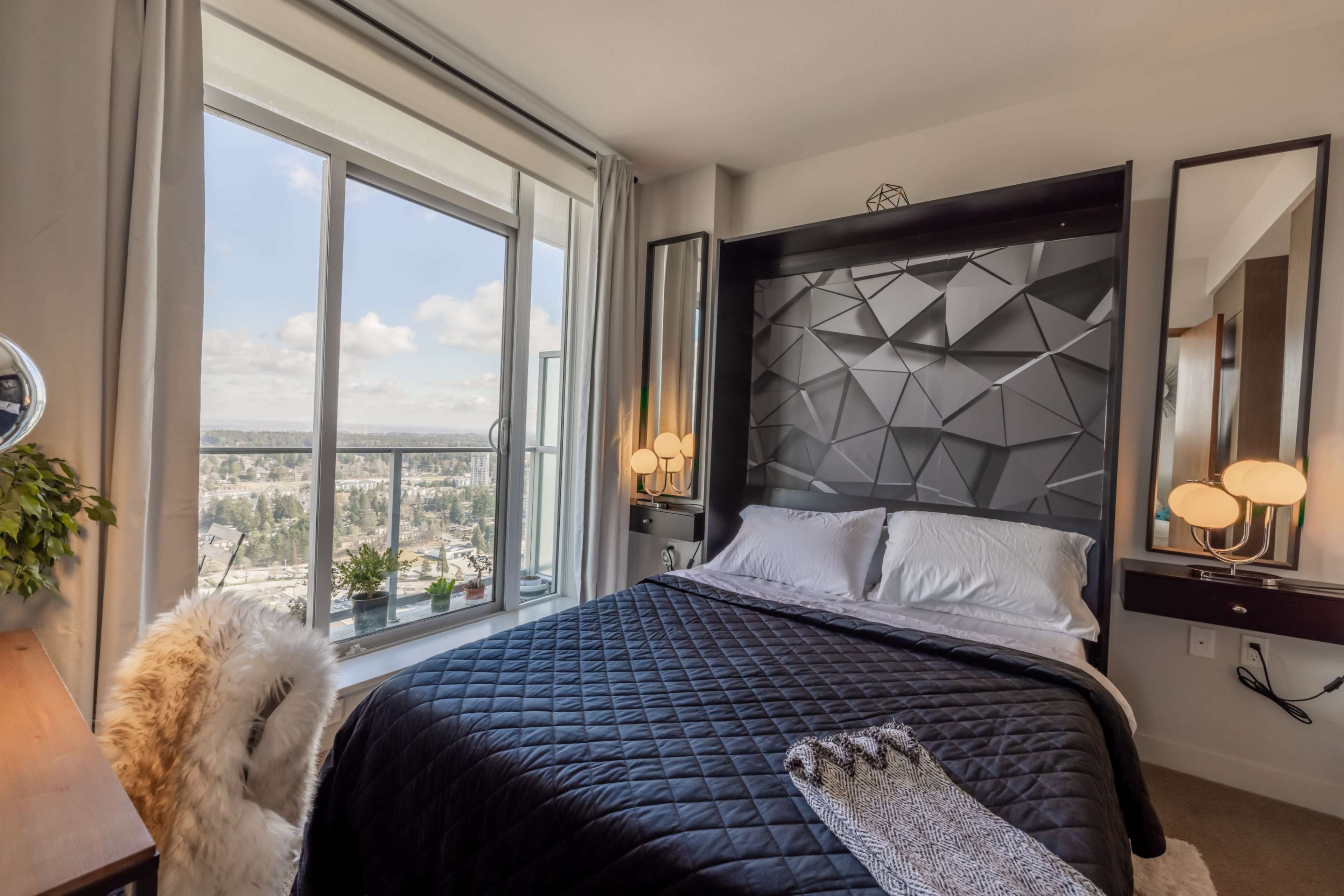 A modern bedroom features a geometric accent wall behind a neatly made bed, with large windows offering a view of the cityscape.