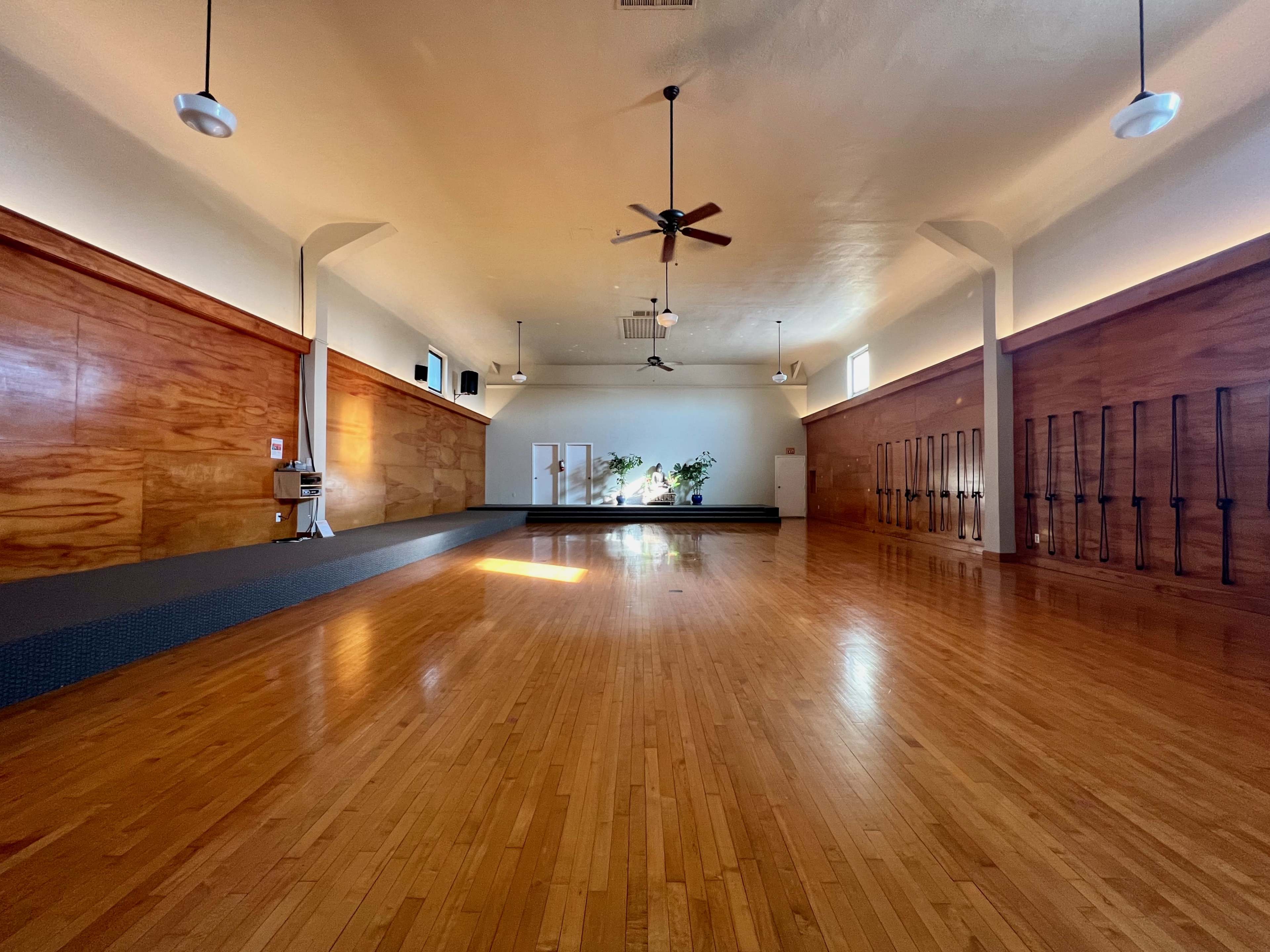 An empty yoga studio with wooden floors, large windows letting in natural light, and wall-mounted yoga props.