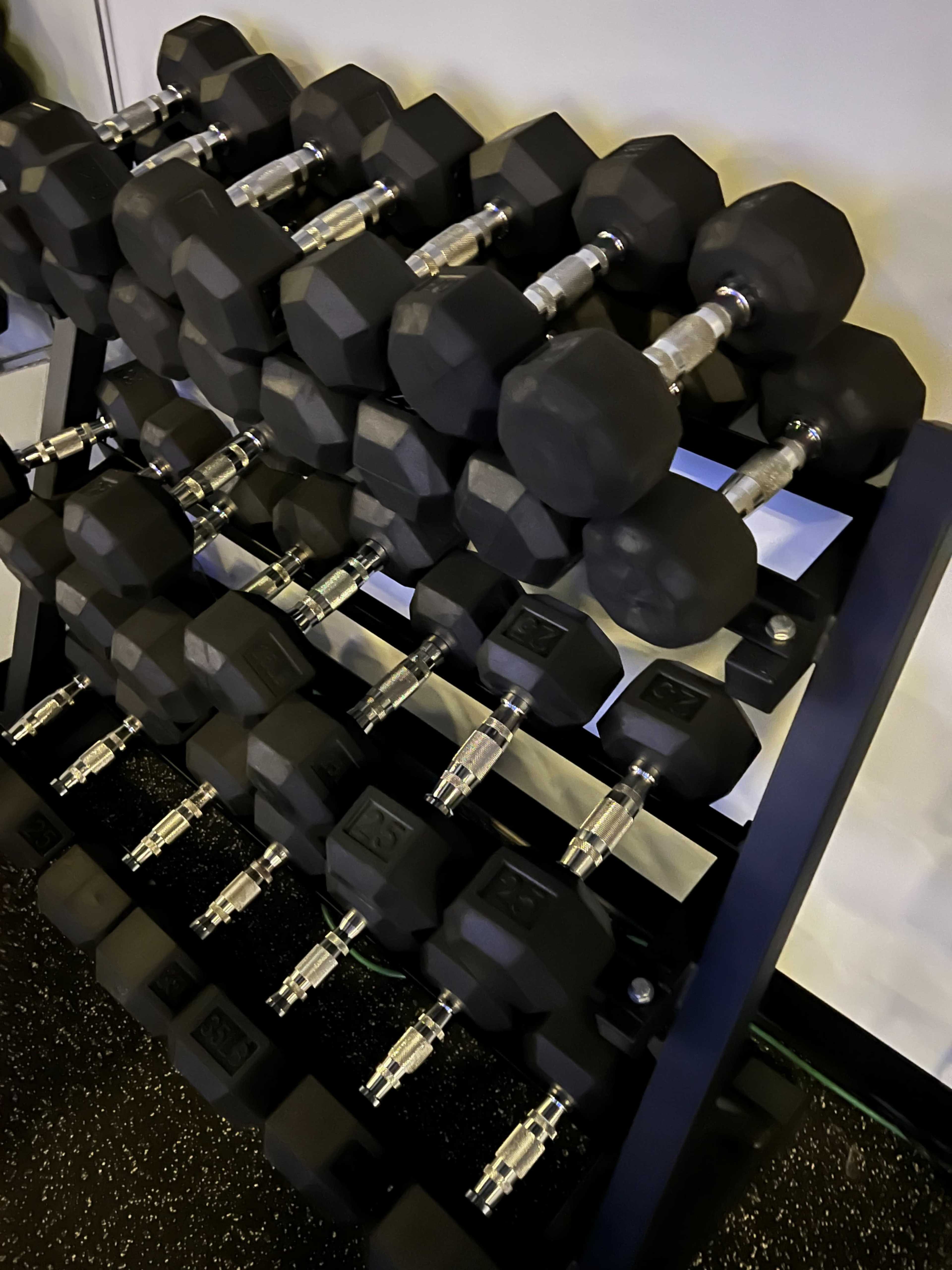 A steel rack holds several rows of black hexagonal dumbbells in a gym setting.