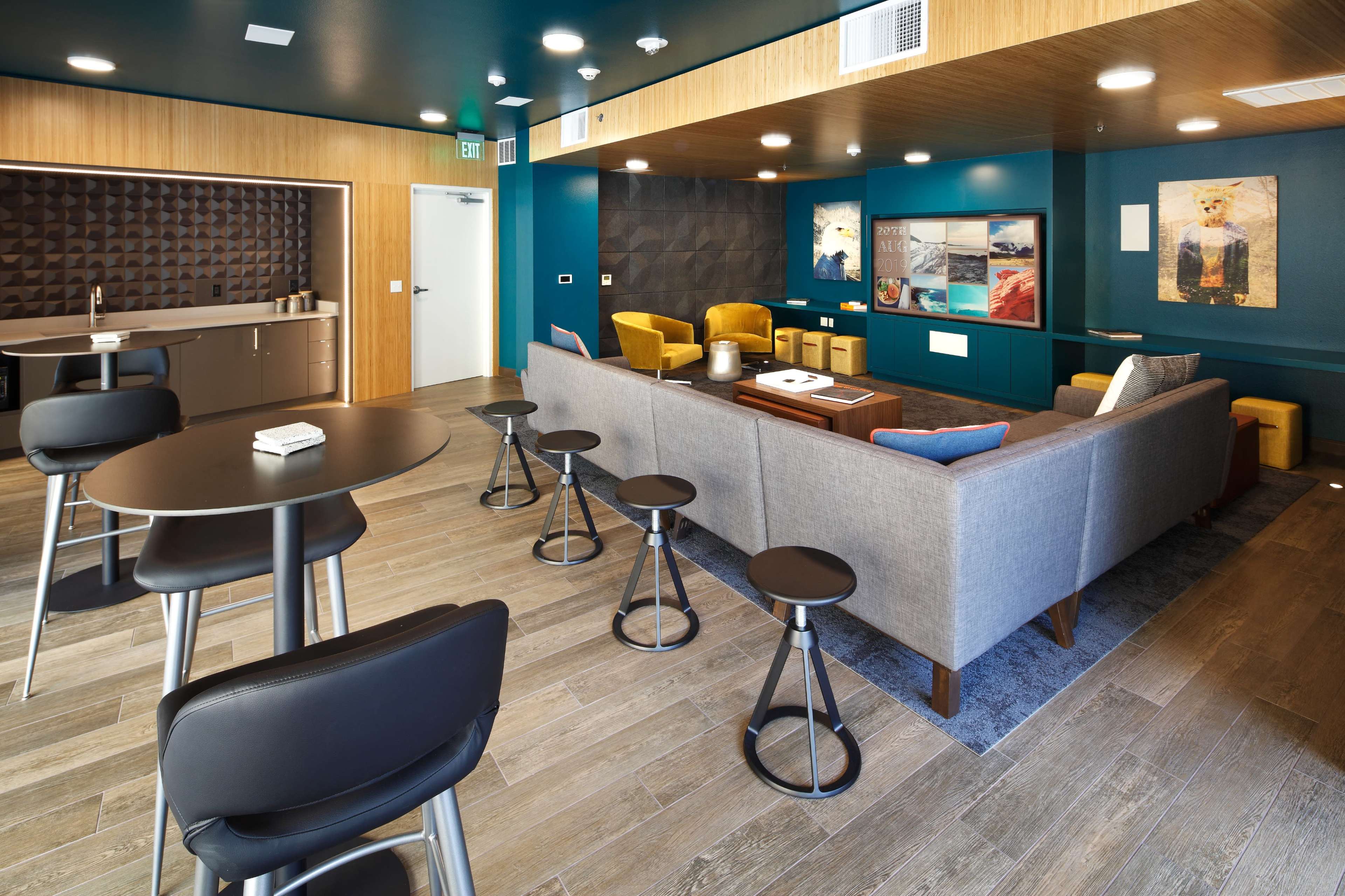 A modern lounge area featuring a gray sectional sofa, yellow armchairs, a coffee table, and a bar counter with high stools against a wooden wall.