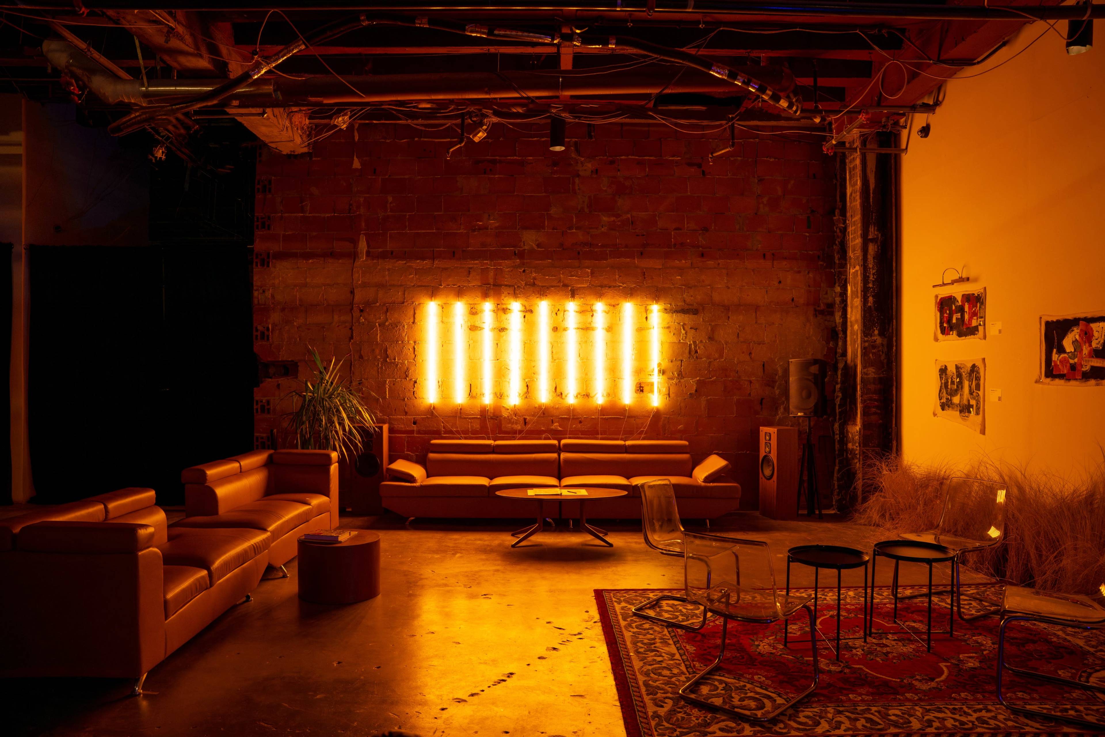 The image shows a stylish urban lounge with leather sofas, a circular coffee table, and illuminated wall art against a brick backdrop.