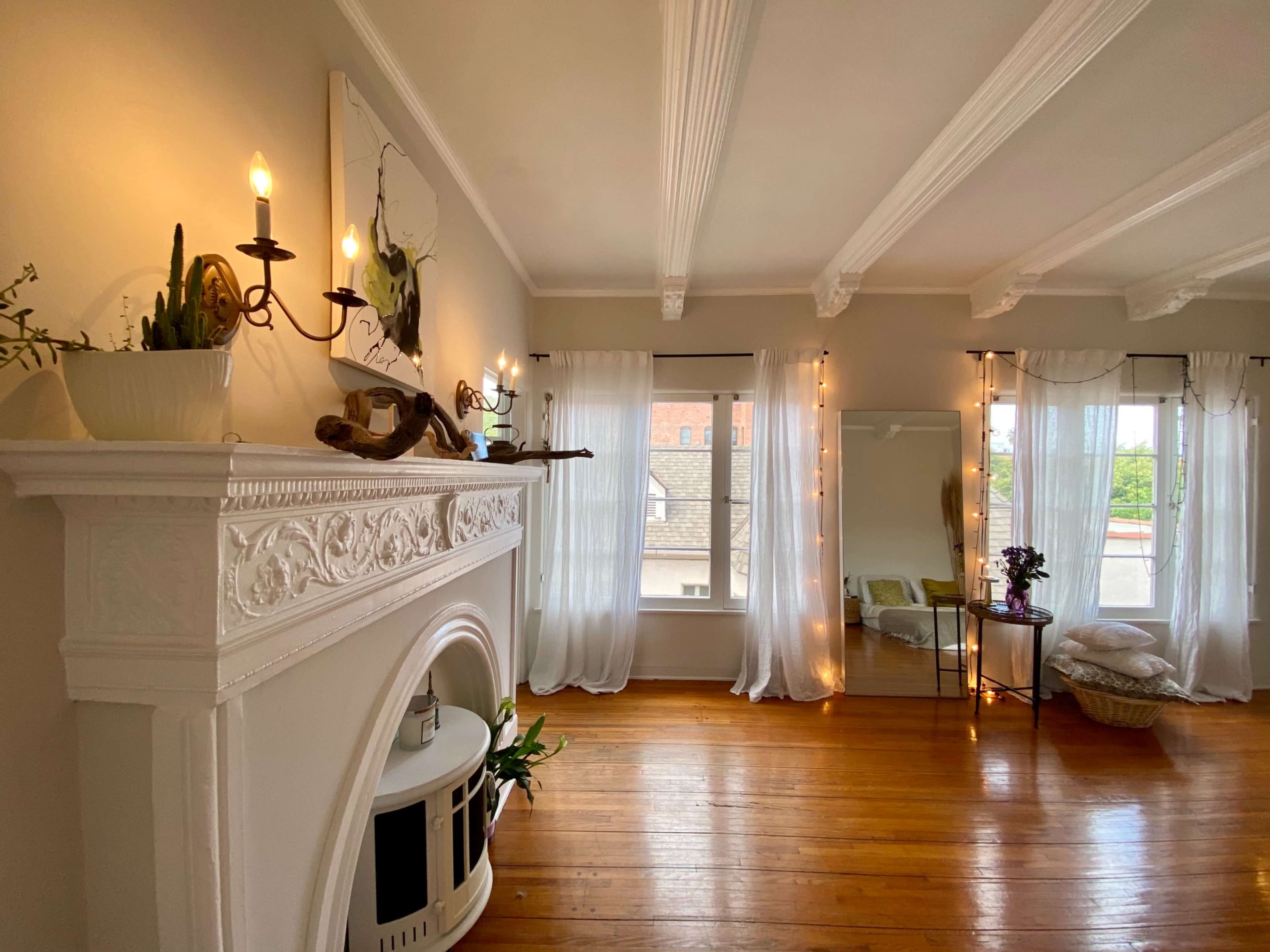 A bright room features a white fireplace with candles, large windows draped in sheer curtains, and a mirror with a small table beside it.