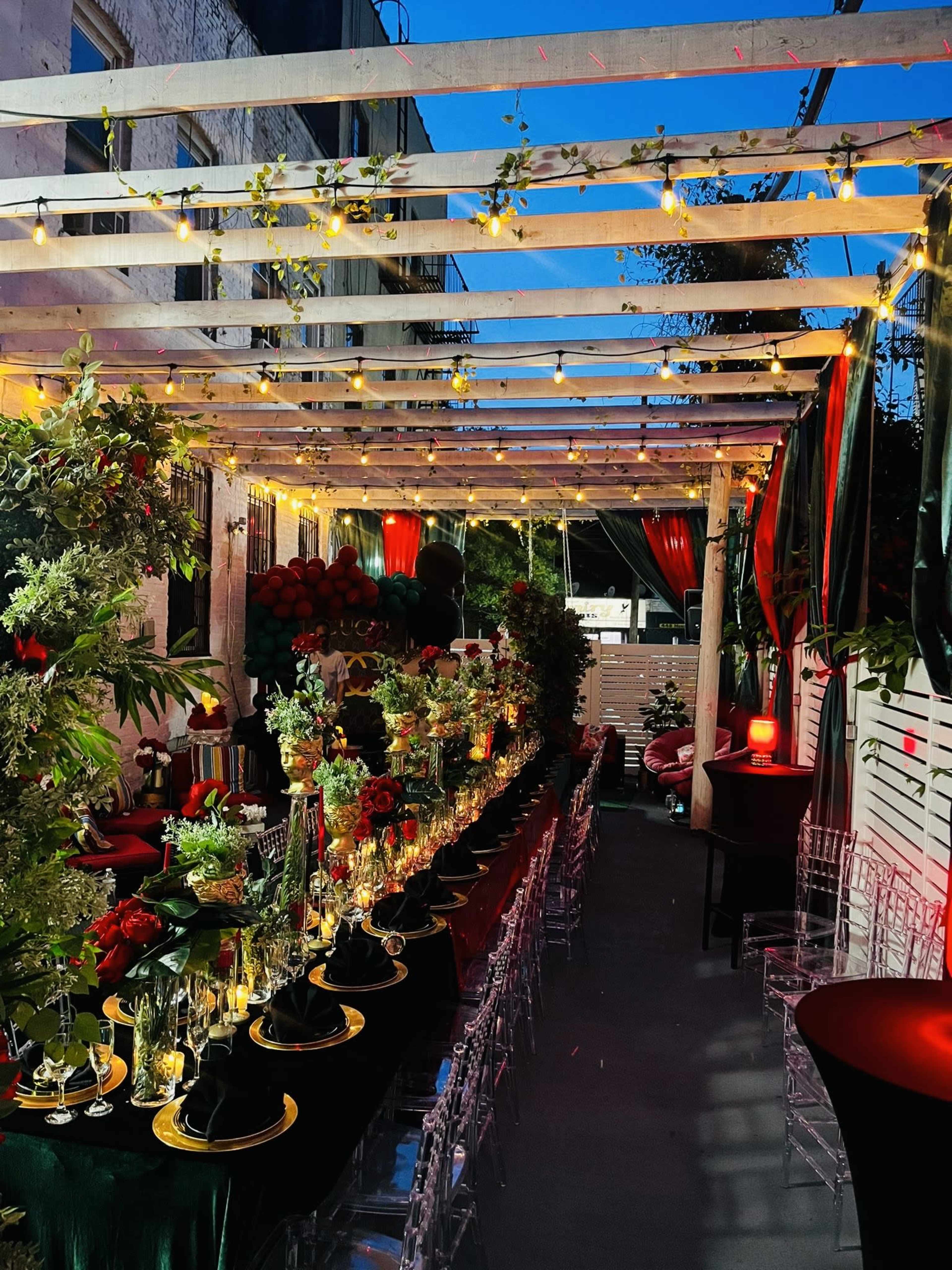 The image shows a beautifully arranged outdoor dining setup under a wooden pergola, adorned with greenery and string lights.