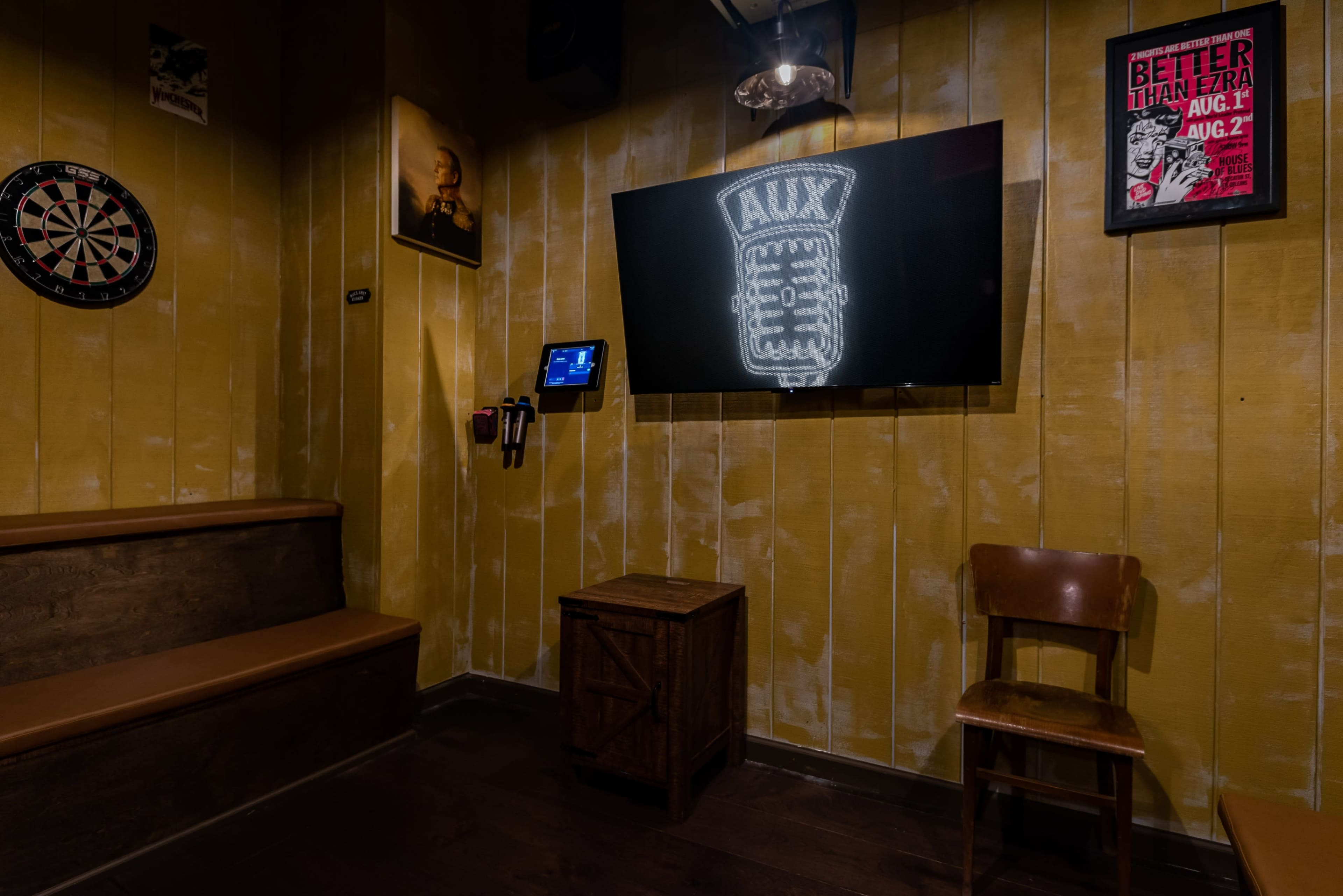 The image shows a cozy room featuring a large screen, a dartboard, a wooden bench, a chair, and framed posters on the walls.