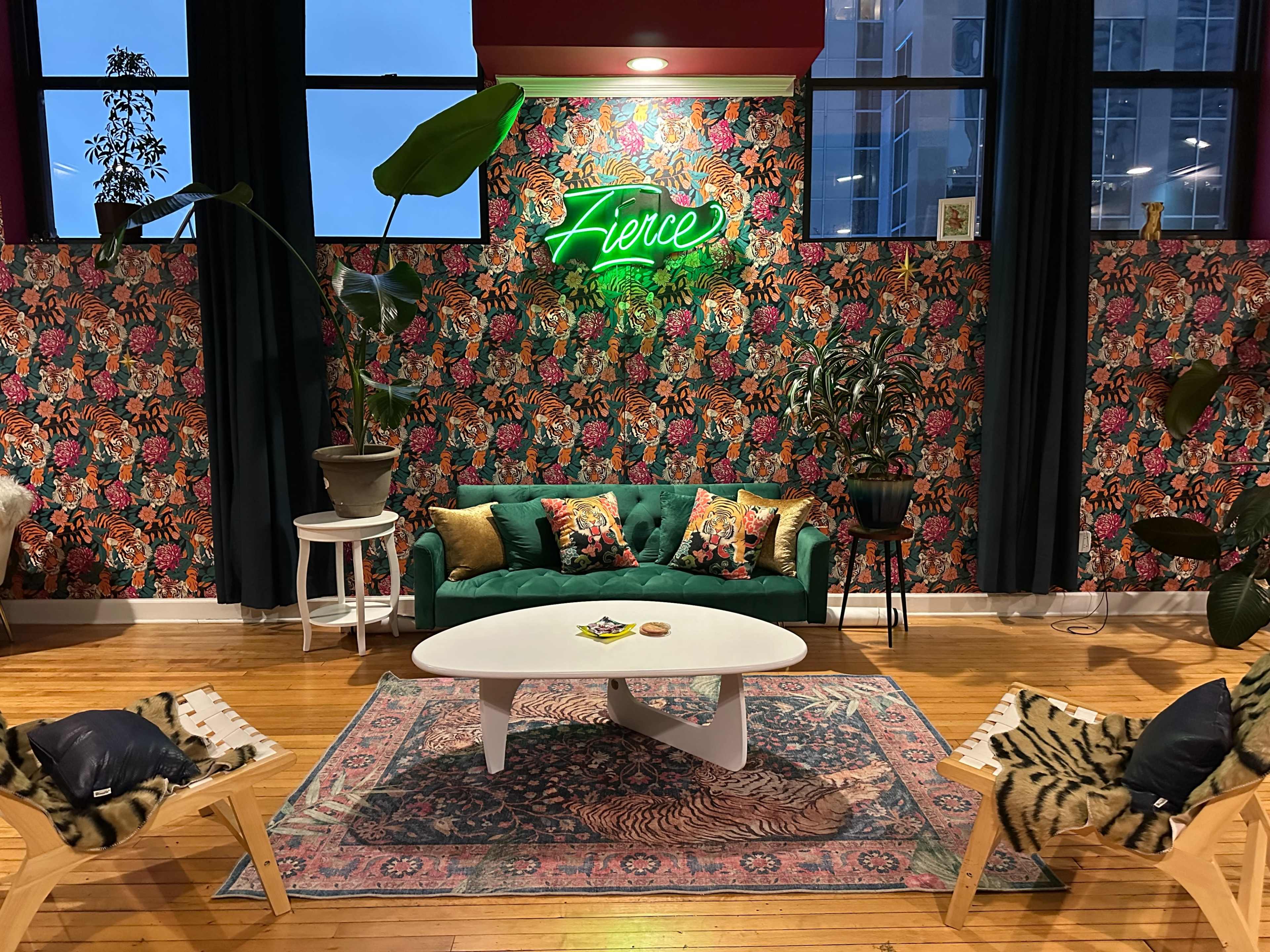 The image shows a vibrant interior featuring a green sofa with decorative pillows, a large floral wallpaper, a neon sign that says "Fierce," and various plants alongside wooden furniture.
