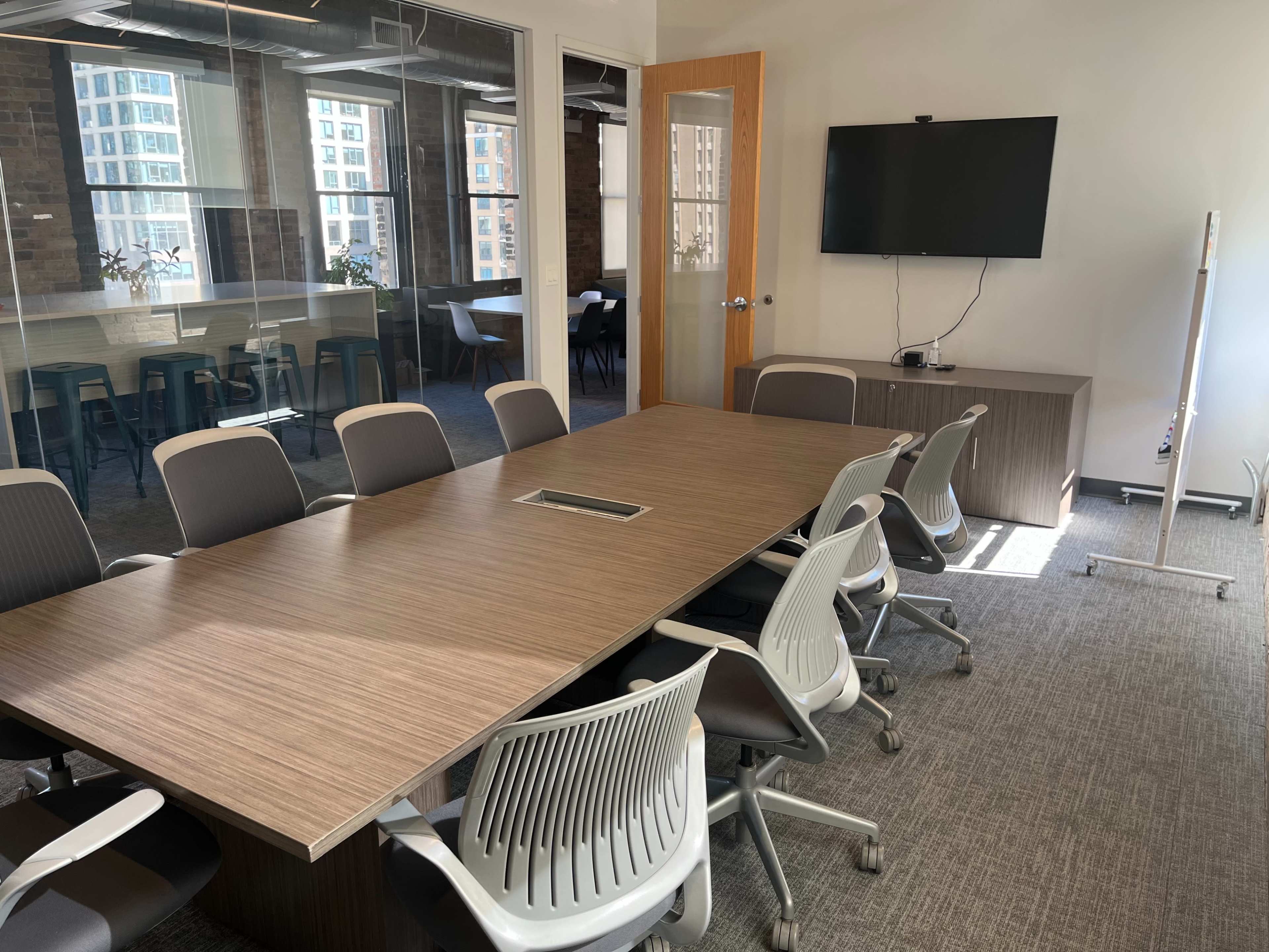 The image shows a modern conference room with a large wooden table surrounded by ergonomic chairs, and a wall-mounted TV.