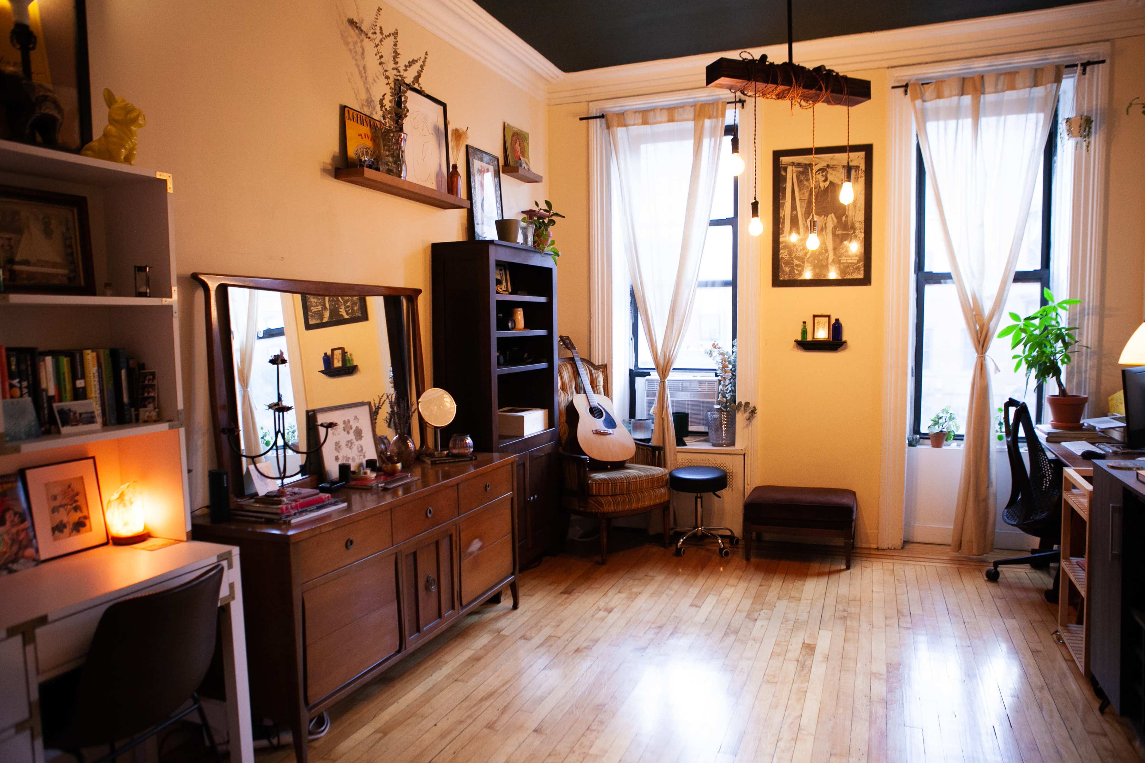 An interior room with wooden floors, a large window, plants, a guitar, and various pieces of furniture arranged throughout.